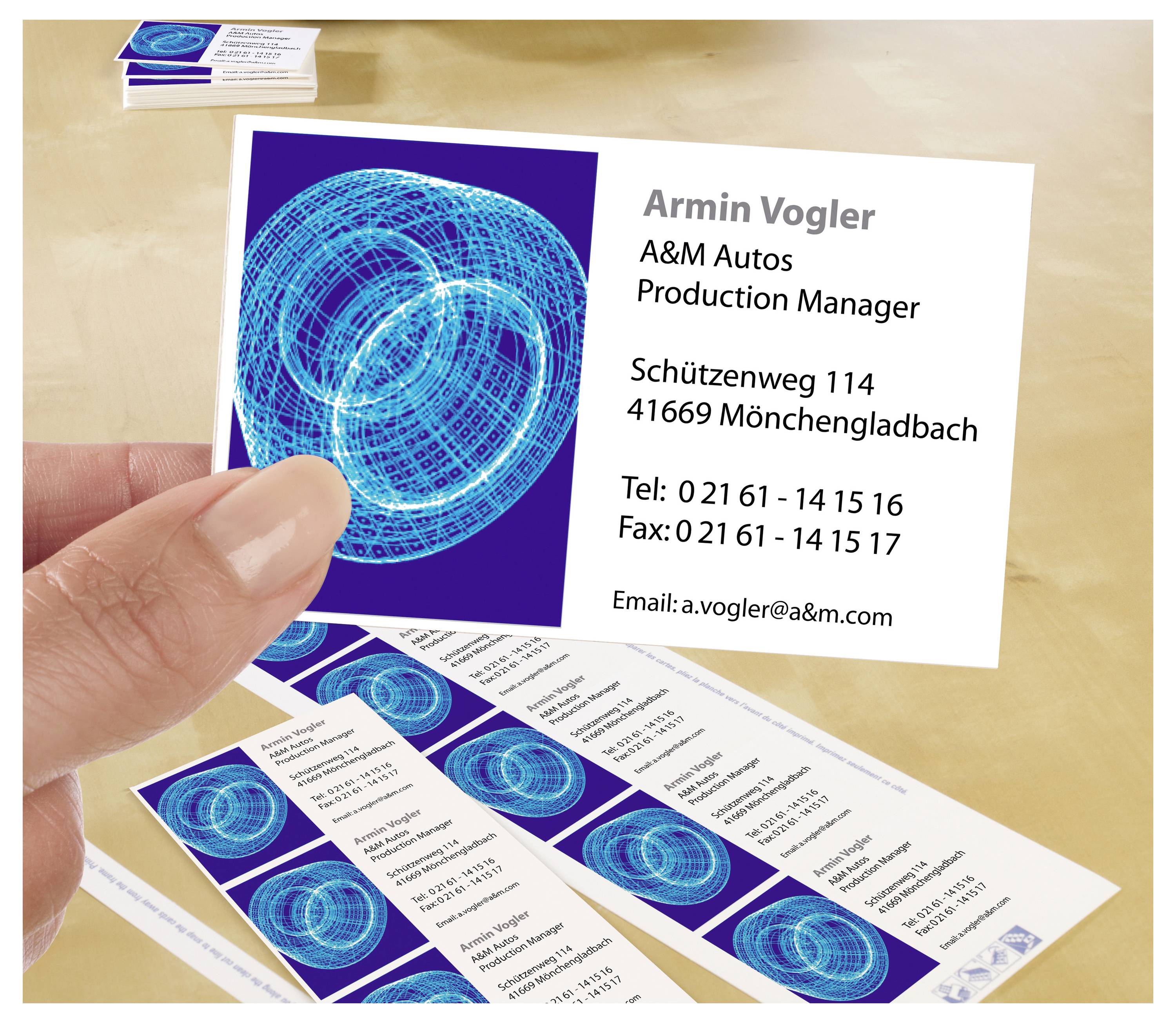 Hand hält eine Visitenkarte von Armin Vogler, A&M Autos, Production Manager, mit Adresse, Telefon, Fax und E-Mail; weitere Karten liegen daneben.