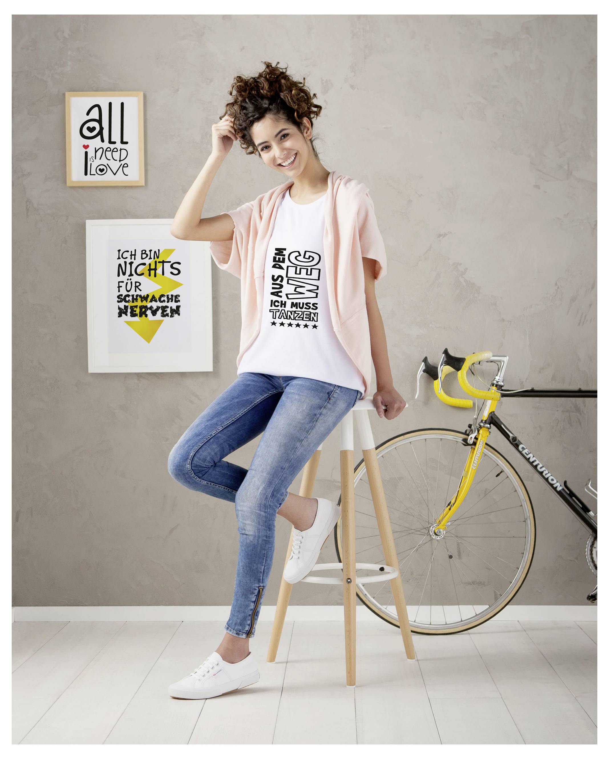 Junge Frau sitzt lächelnd auf einem Hocker neben einem gelben Rennrad; im Hintergrund hängen zwei gerahmte Poster mit englischer und deutscher Typografie.