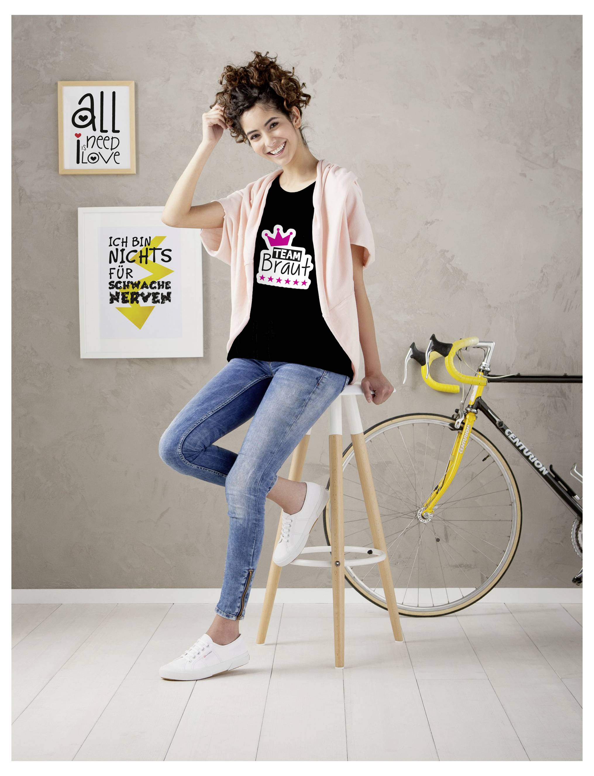 Junge Frau sitzt lächelnd auf einem Hocker vor einer Wand mit Motiven; auf ihrem T‑Shirt steht 'TEAM BRAUT', neben ihr lehnt ein gelbes Fahrrad.