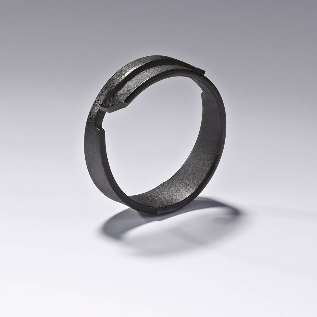 Ein schwarzer, kreisförmiger Metallring liegt auf einer grauen Oberfläche. Der Ring hat eine kleine, eingekerbte Öffnung an der Seite.