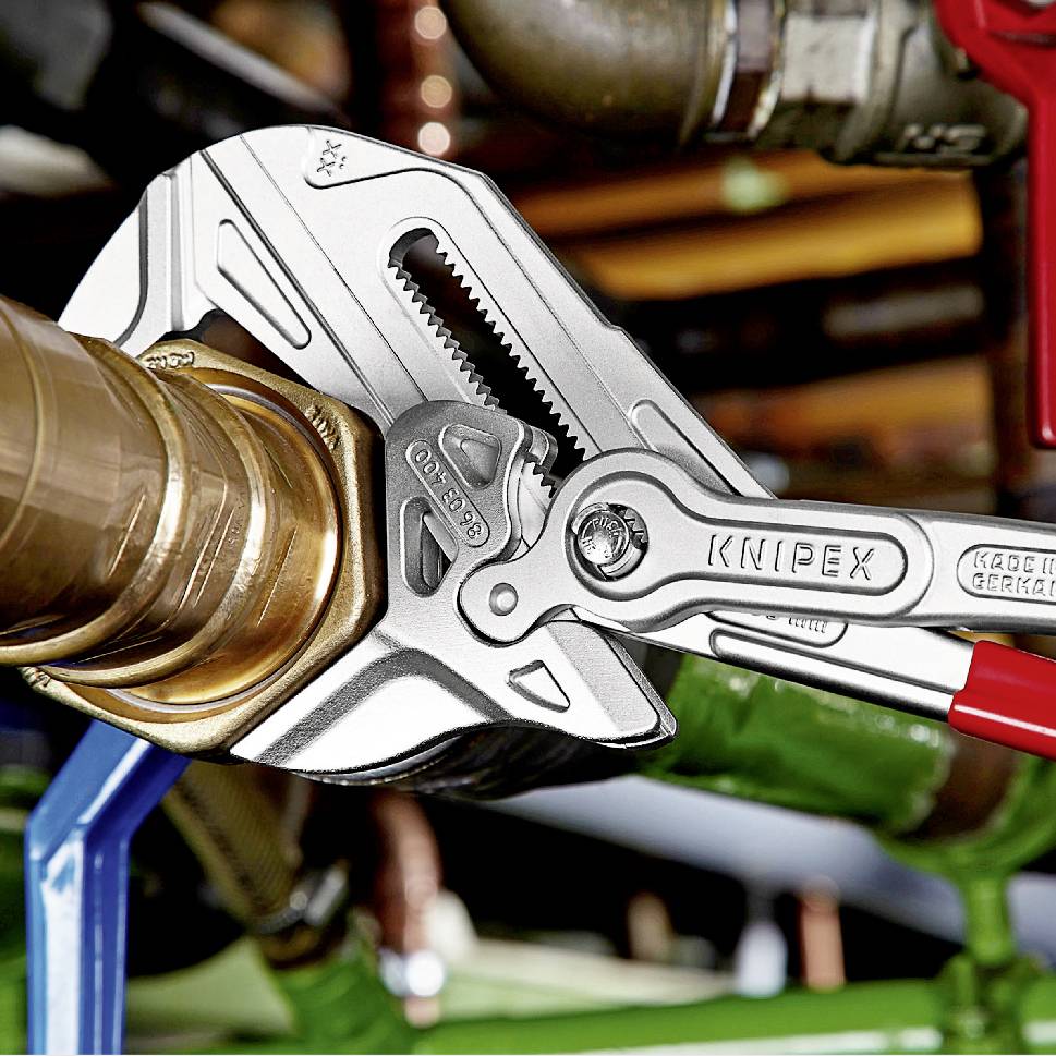 Ein verstellbarer Knipex-Rohrzangenschlüssel greift eine Metallverschraubung an einem Rohr. Werkzeuge und Rohre im Hintergrund.