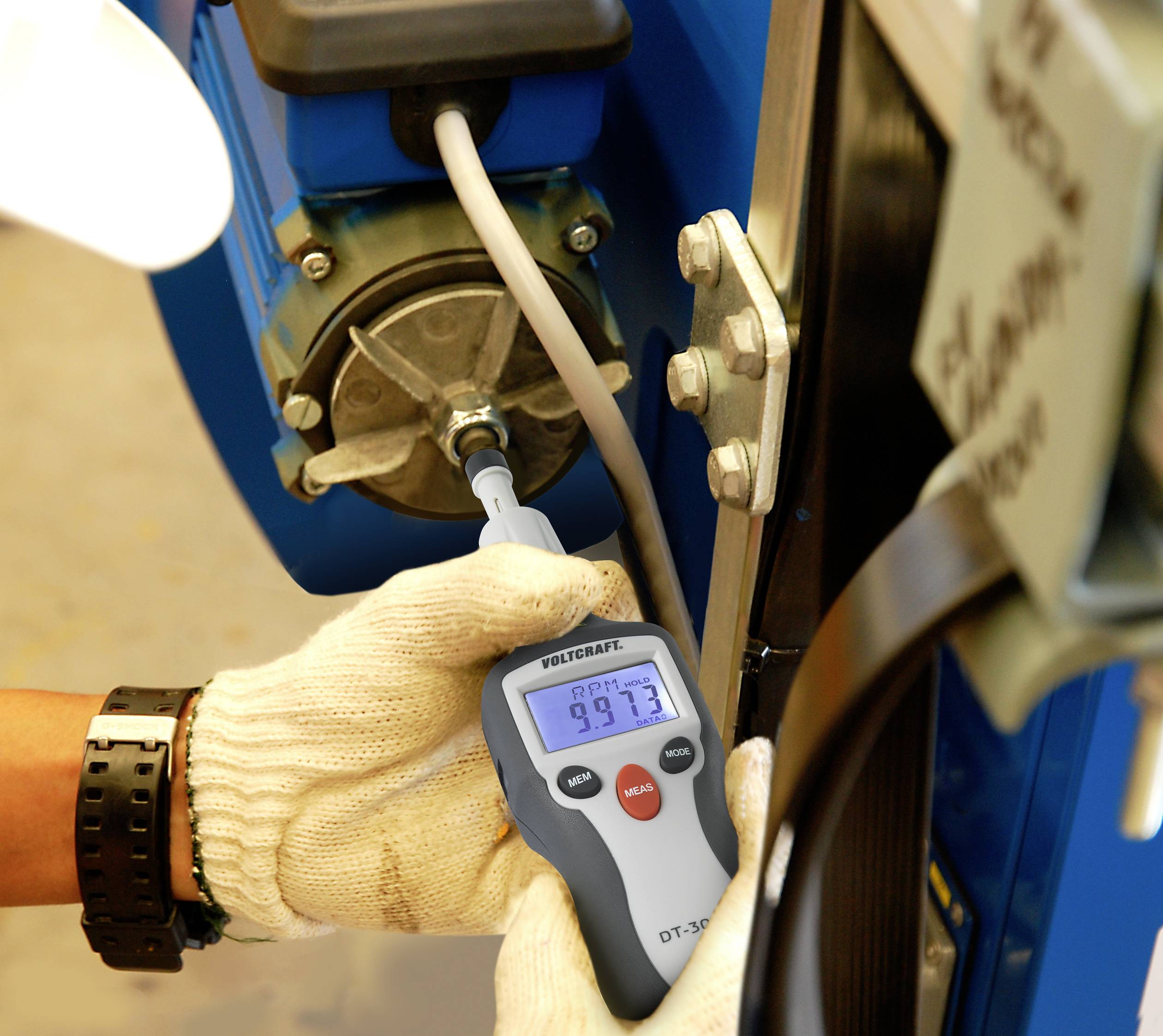 Eine Person misst mit einem Handgerät die Drehzahl eines Motors. Das Display zeigt '9973'. Die Hände tragen weiße Handschuhe.