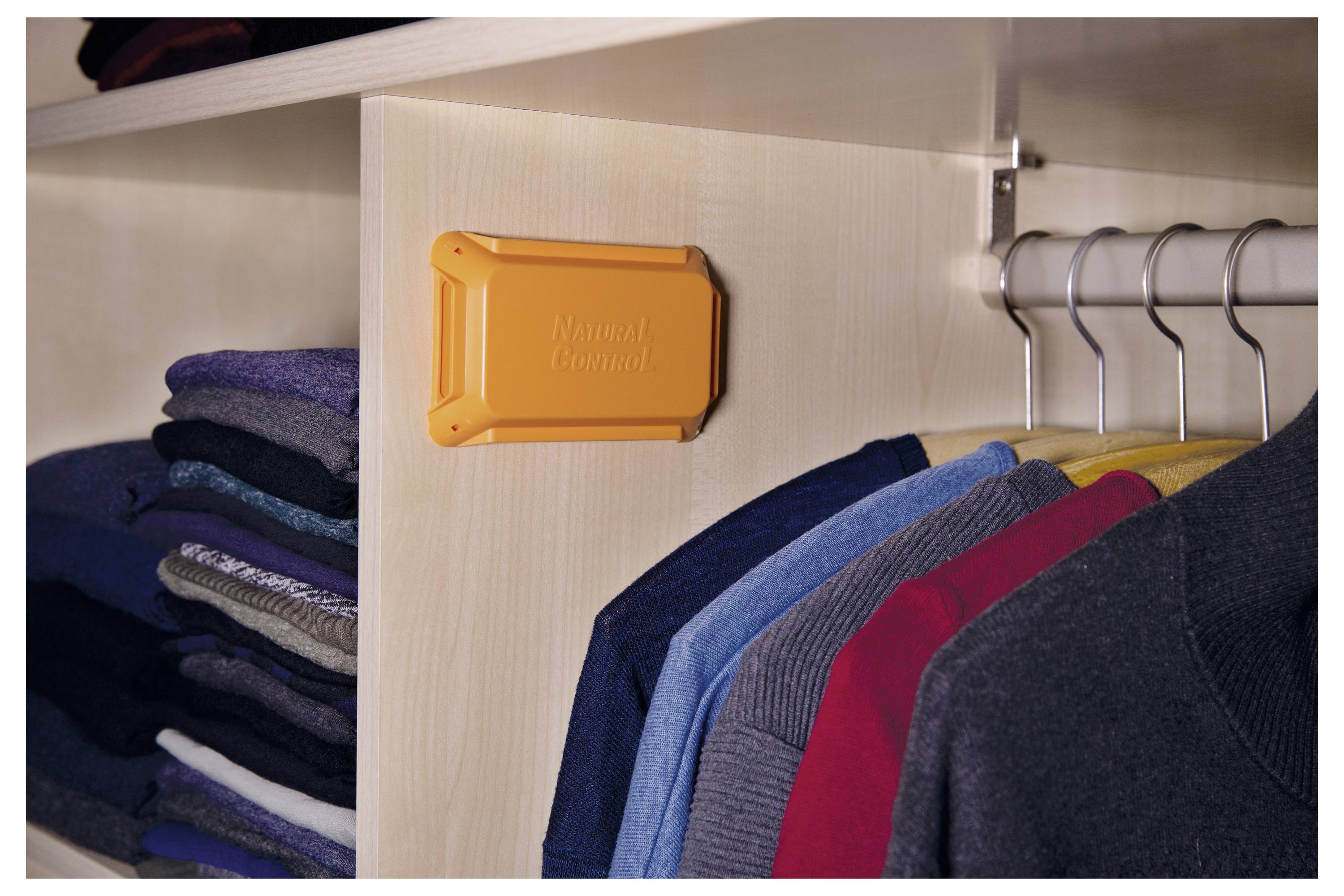 Kleidungsstücke hängen in einem Kleiderschrank, an dessen Seite ein orangefarbenes Plastikgerät befestigt ist. Das Gerät trägt die Aufschrift 