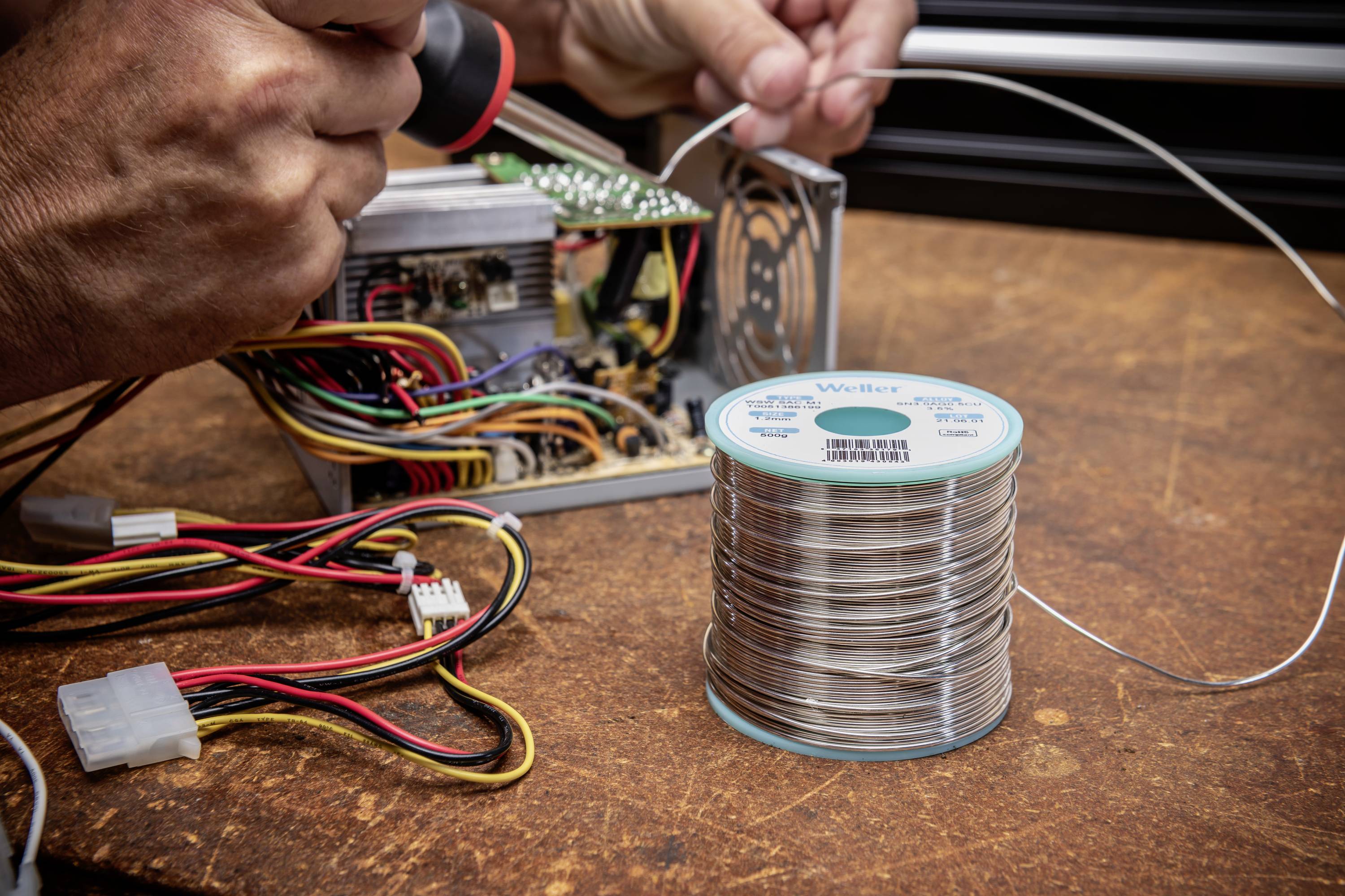 Eine Person lötet Kabel an ein elektronisches Gerät. Ein Lötdraht steht im Vordergrund auf einem Holztisch.