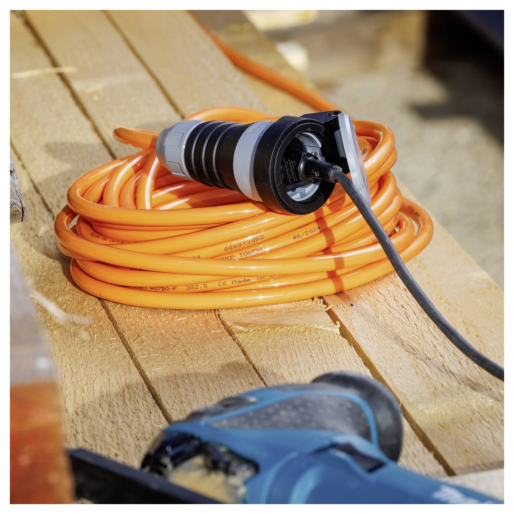 Aufgewickeltes orangefarbenes Verlängerungskabel mit schwarzem Stecker auf einer Holzoberfläche, mit einem blau sichtbaren Elektrowerkzeug im Vordergrund.