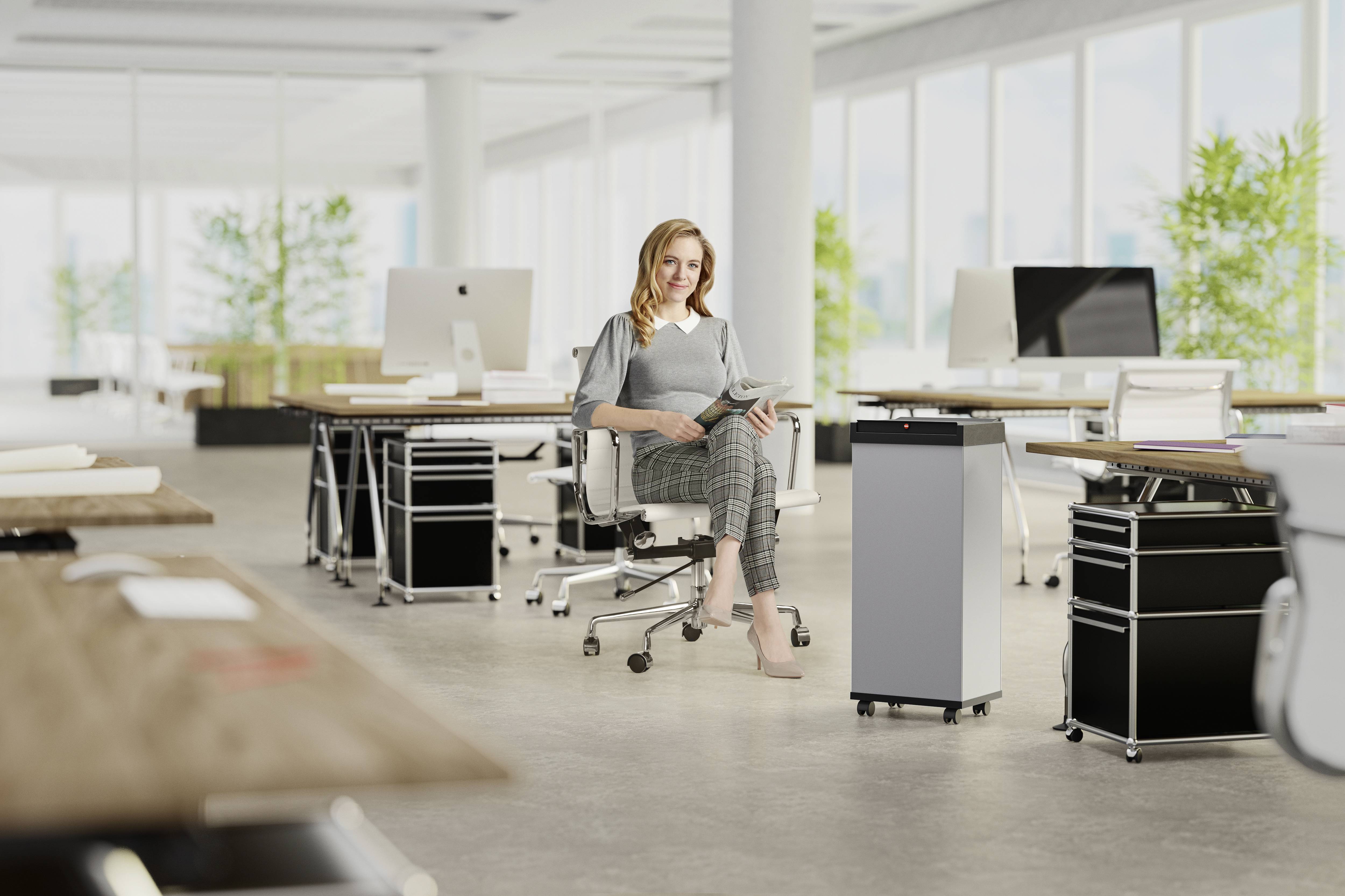 Eine Frau sitzt mit einem Tablet in einem modernen Büro mit mehreren Arbeitsplätzen. Große Fenster im Hintergrund lassen viel Licht herein.