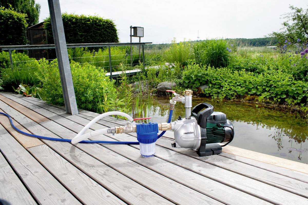 Eine Wasserpumpe mit Schläuchen steht auf einer Holzterrasse neben einem Teich, umgeben von üppiger Vegetation im Hintergrund.