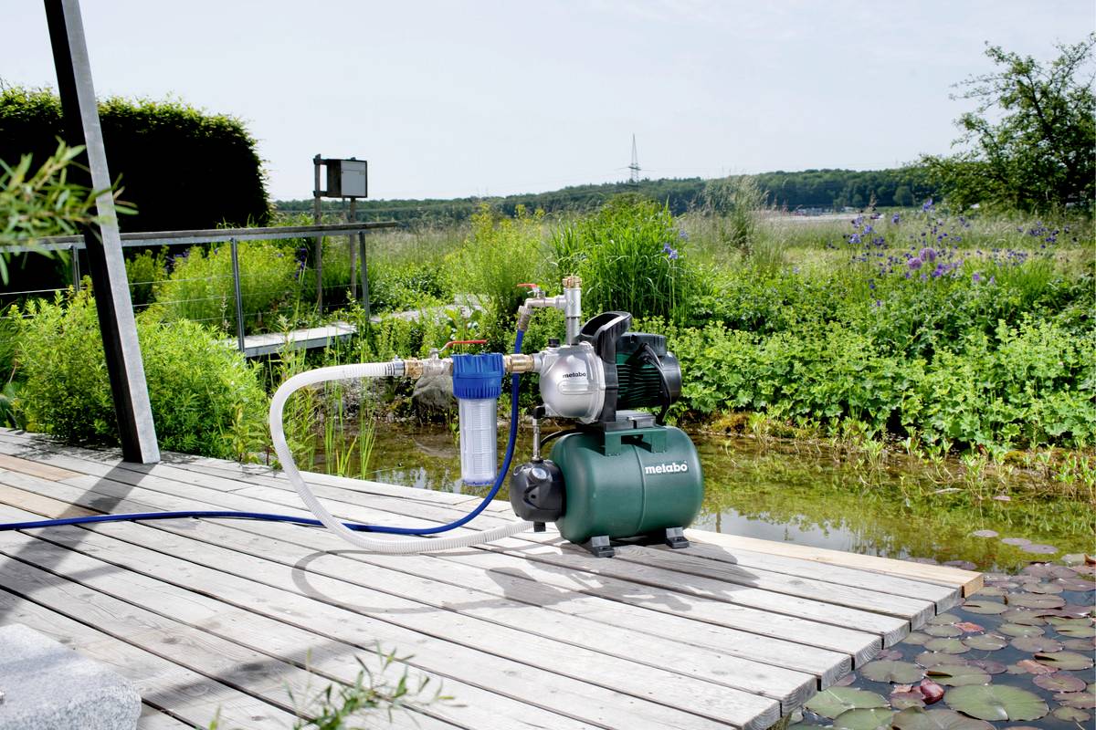 Eine Wasserpumpe auf einer Holzterrasse vor einem Teich, umgeben von Pflanzen. Im Hintergrund ist eine Landschaft mit Bäumen sichtbar.