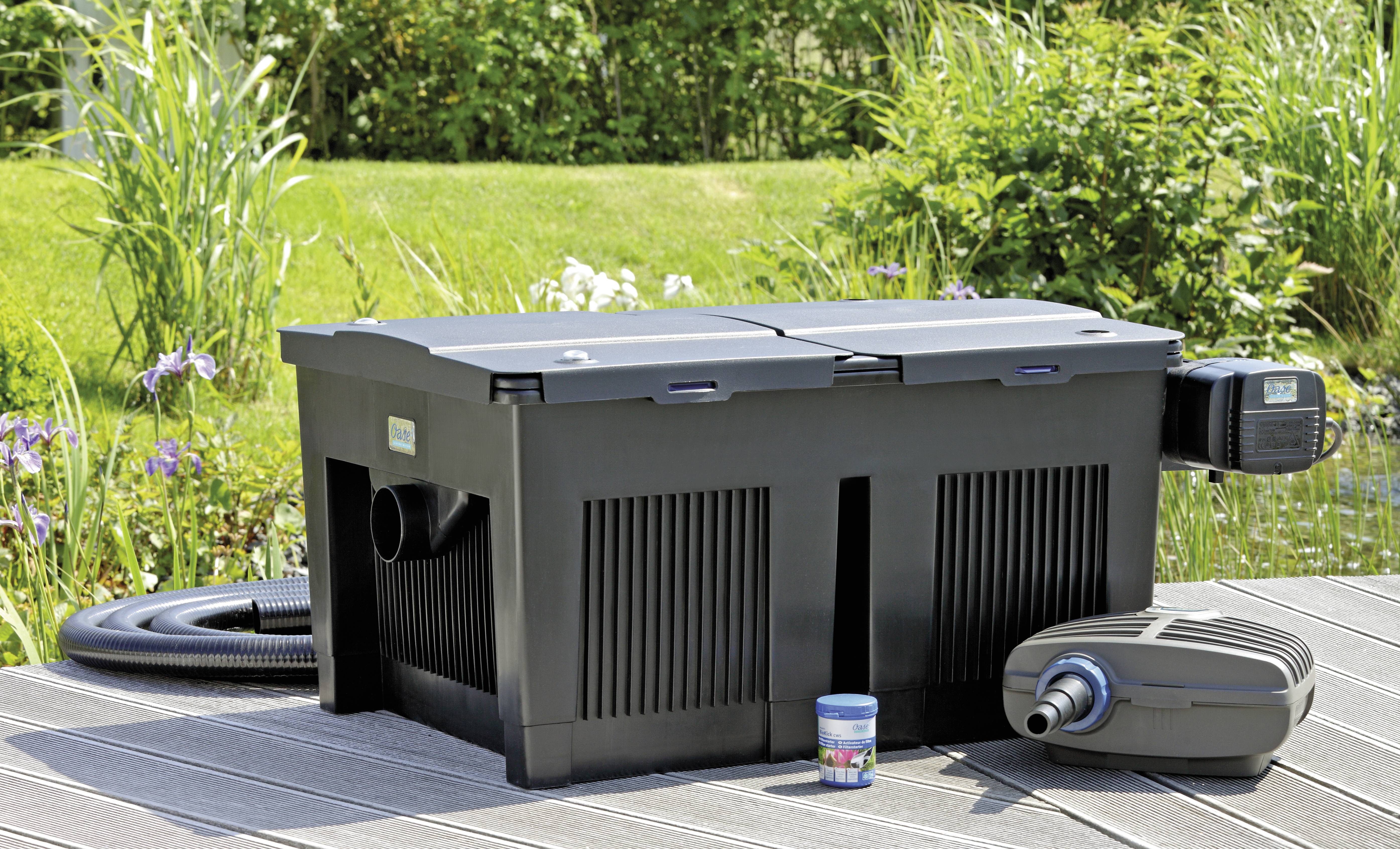 Ein schwarzes Teichfilterset mit Schläuchen und Zubehör steht auf einer Holzterrasse mit Grünanlage im Hintergrund.