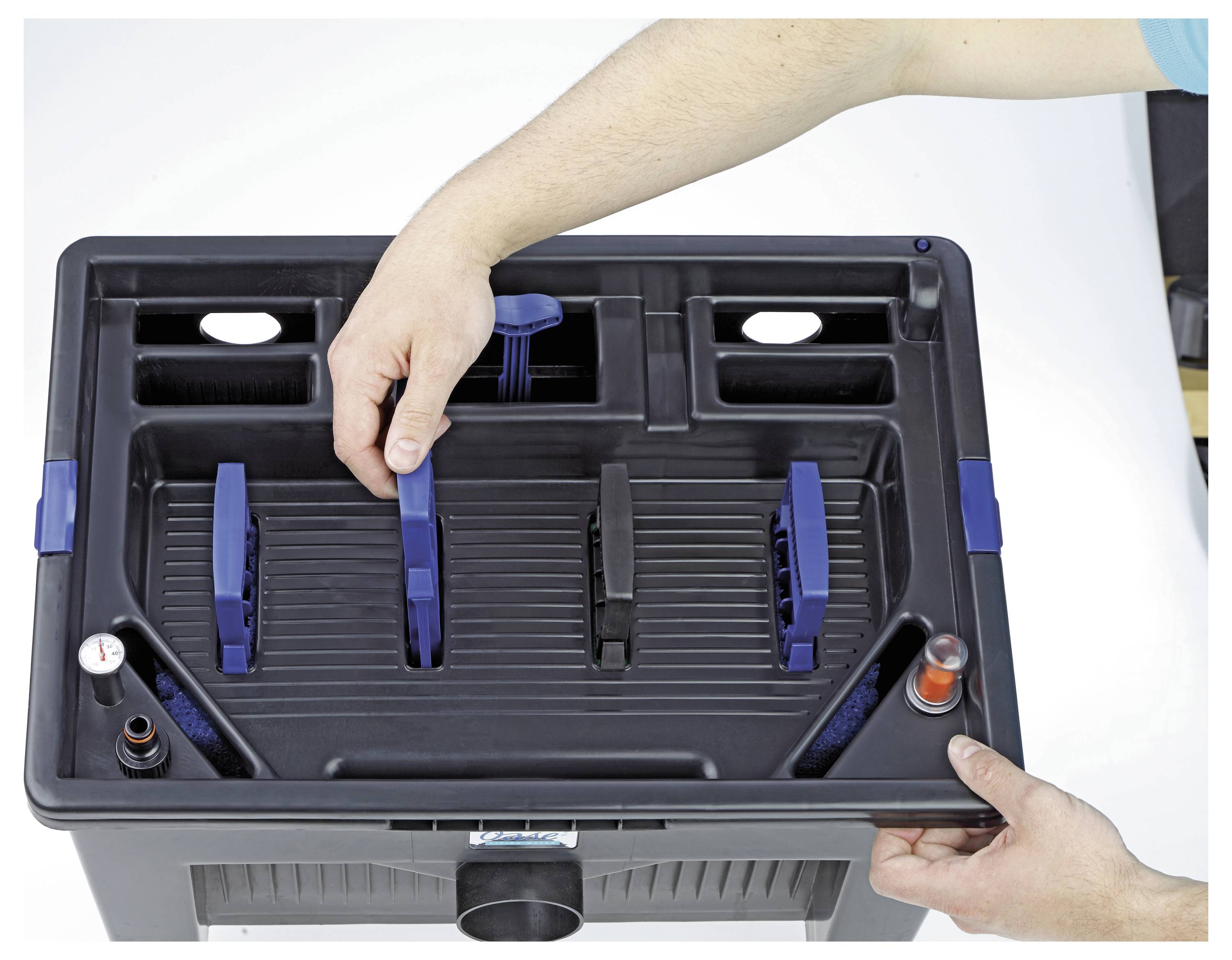 Eine Person justiert eine Komponente in einer schwarzen Plastikbox, die zur Reinigung oder Wartung verwendet wird und blaue Griffe und Fächer aufweist.