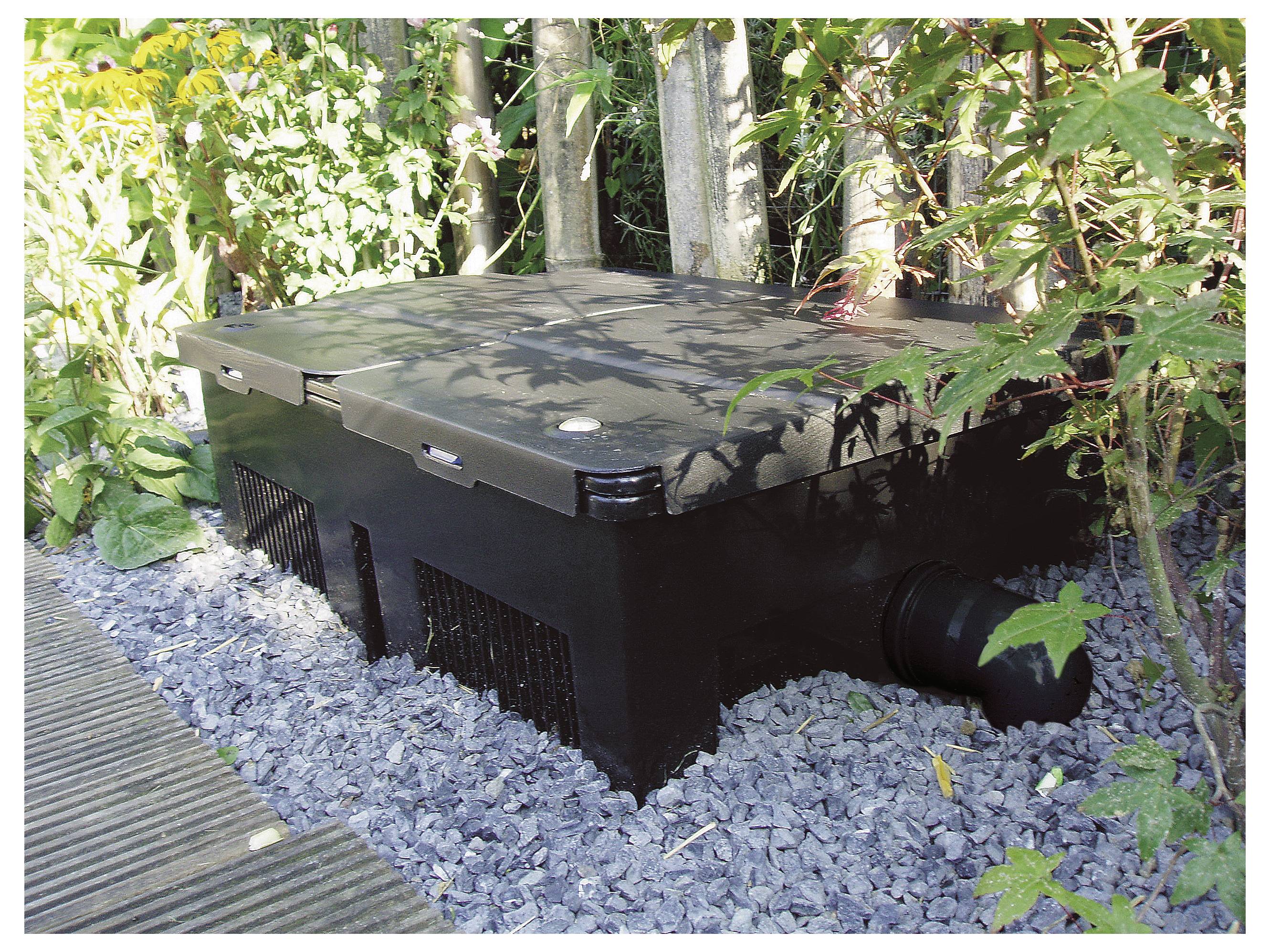 Eine schwarze Teichfilterbox, umgeben von Grünpflanzen und auf Kies platziert, neben einer Holzterrasse, mit sichtbaren Rohren an ihrer Seite.