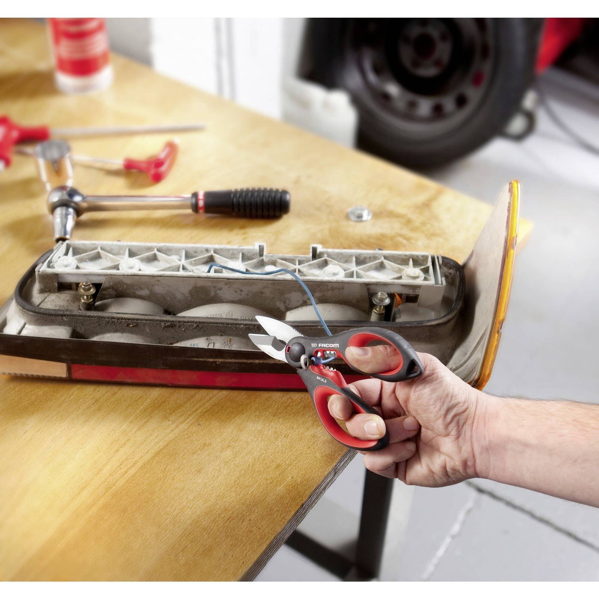 Eine Hand hält eine rote Zange und schneidet ein blaues Kabel, das mit einer Autolampe auf einem Holztisch verbunden ist. Im Hintergrund sind Werkzeuge zu sehen.