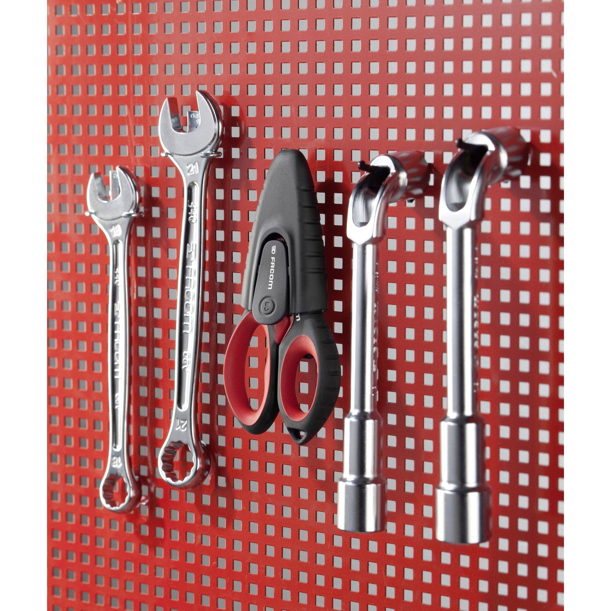 Schraubenschlüssel, Schere und Stecknüsse hängen ordentlich an einer roten Lochwand, in einer Werkstatt oder Garage.