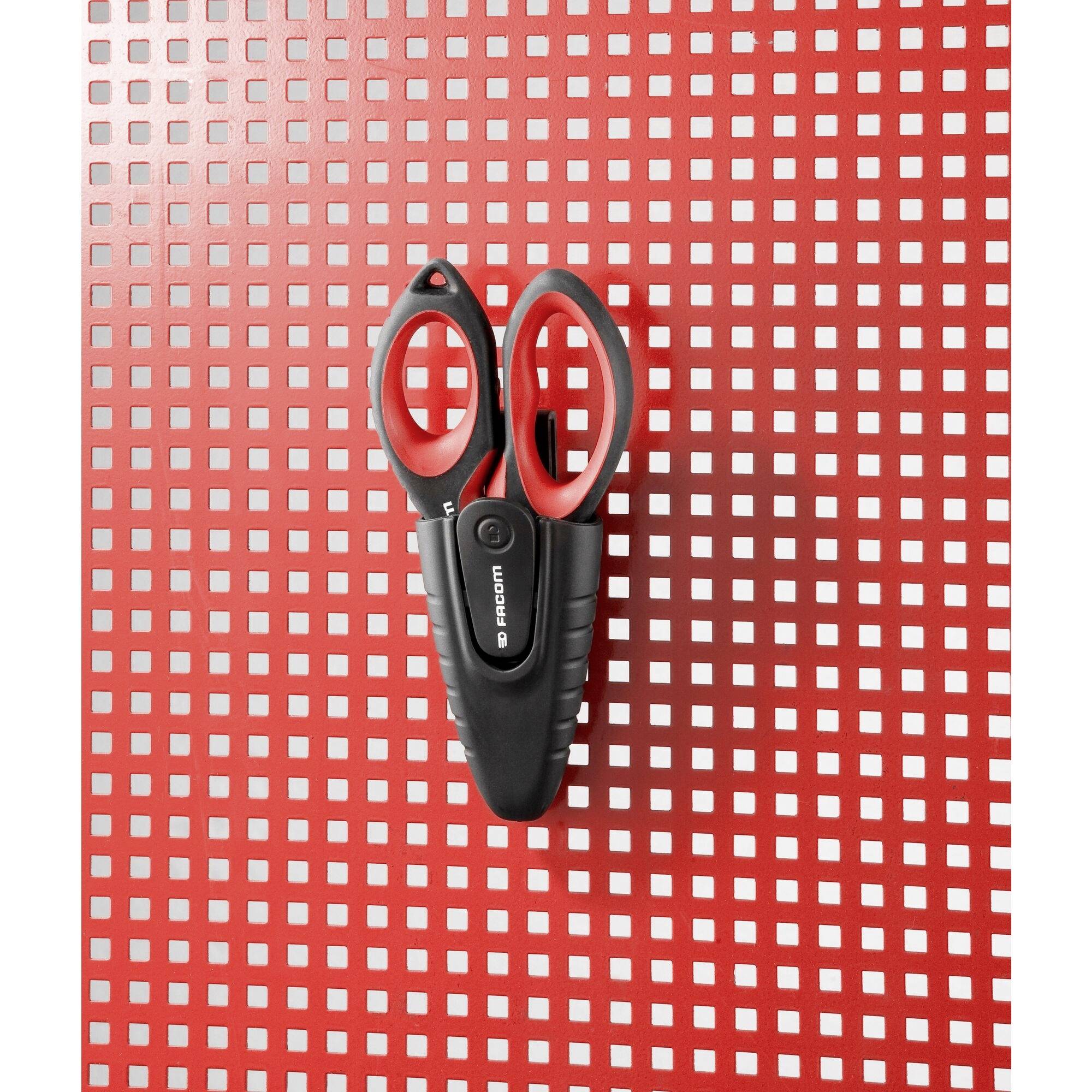 Eine schwarze und rote Schere hängt an einer roten Lochwand mit quadratischem Muster.