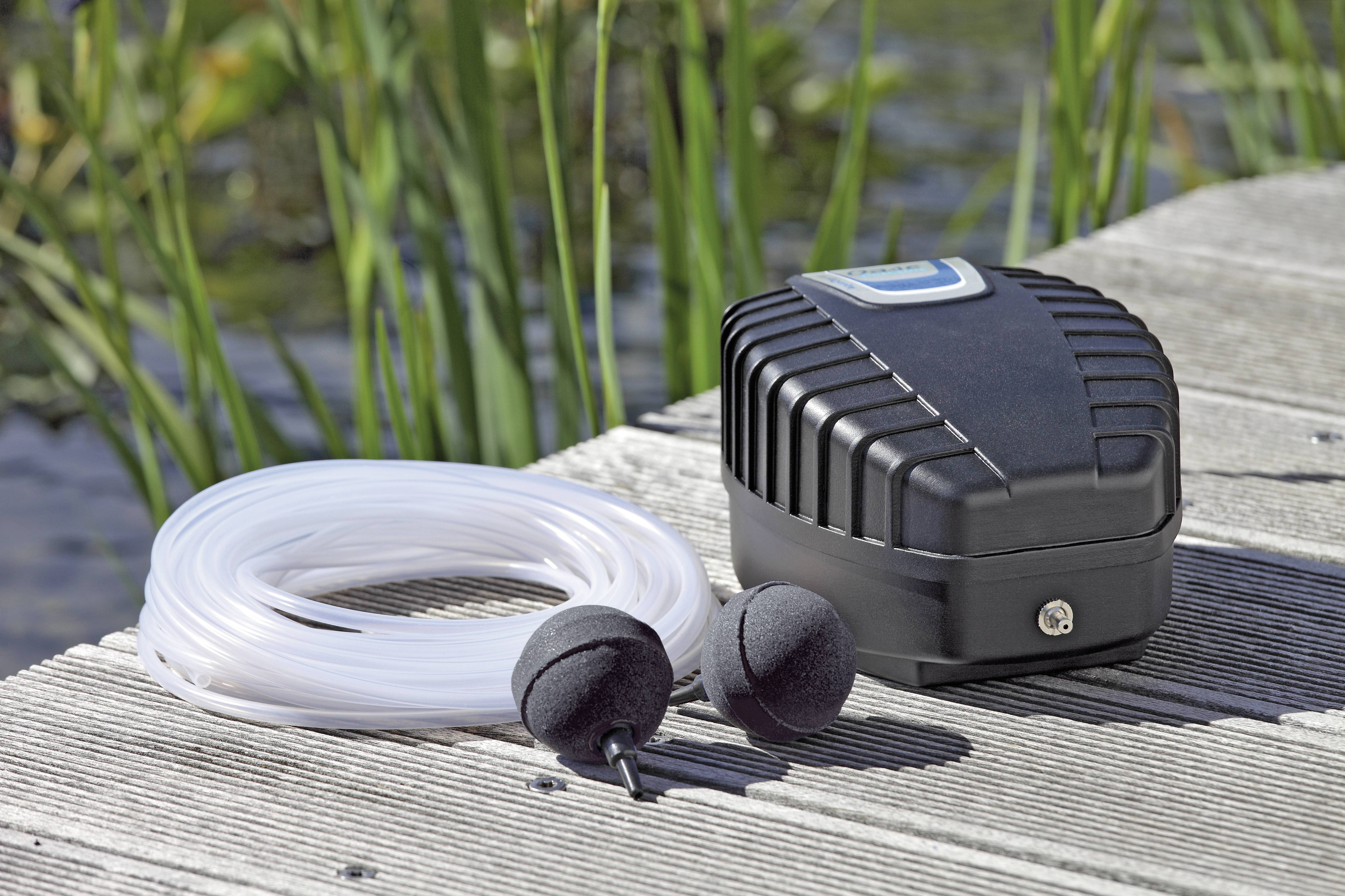 Eine schwarze Luftpumpe liegt auf einem Holzdock neben aufgerollten Schläuchen und zwei runden Diffusoren, mit hohem Gras und Wasser im Hintergrund.