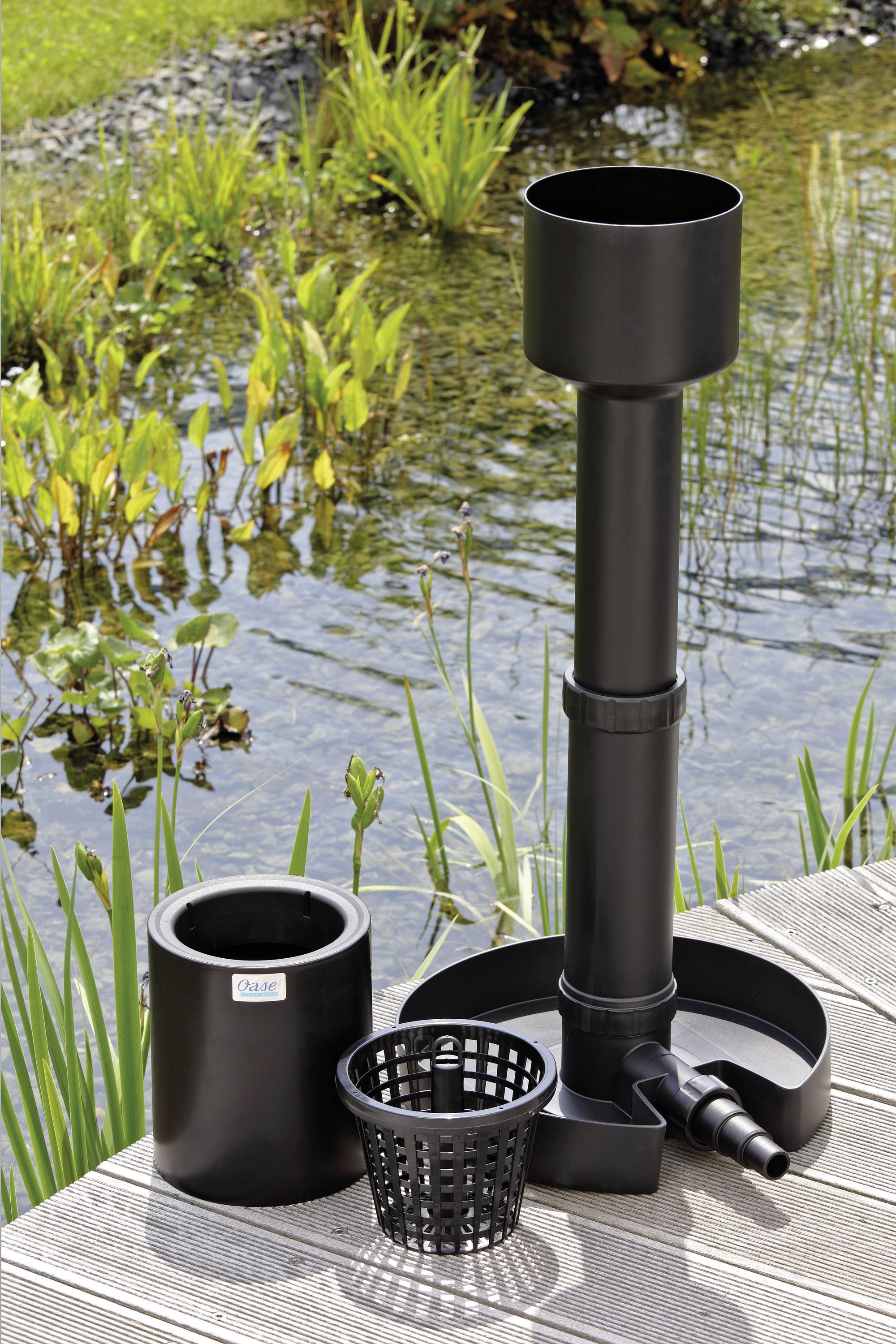 Ein Teichfiltersystem, das neben einem Teich mit Pflanzen aufgestellt ist. Das System umfasst einen hohen schwarzen Filter, einen kleineren runden Behälter und einen Korb.