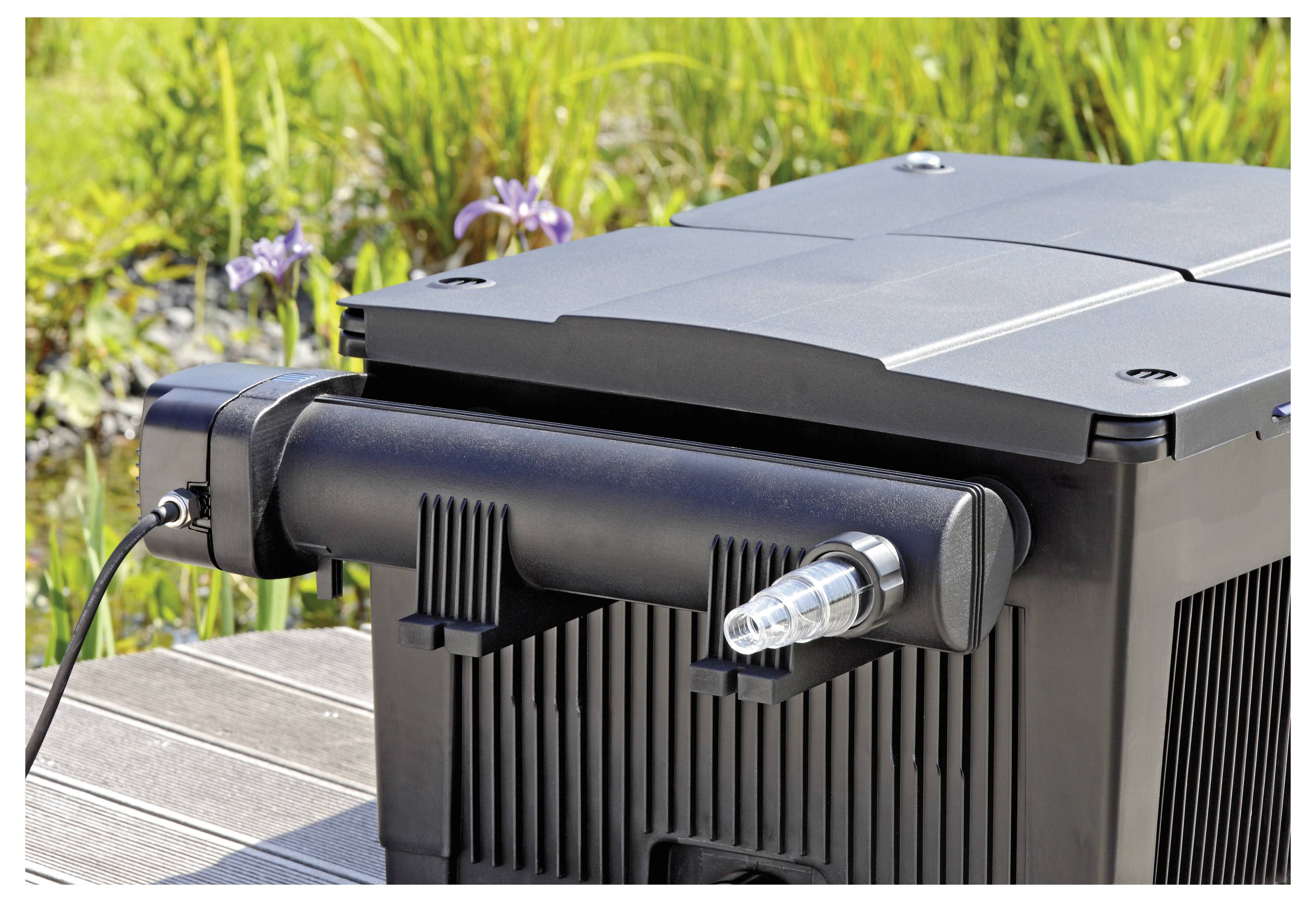 Eine schwarze Kunststoff-Filterbox mit gerippter Oberfläche, versehen mit einem Schlauchanschluss, ist im Freien auf einer Holzoberfläche mit Grünpflanzen im Hintergrund platziert.