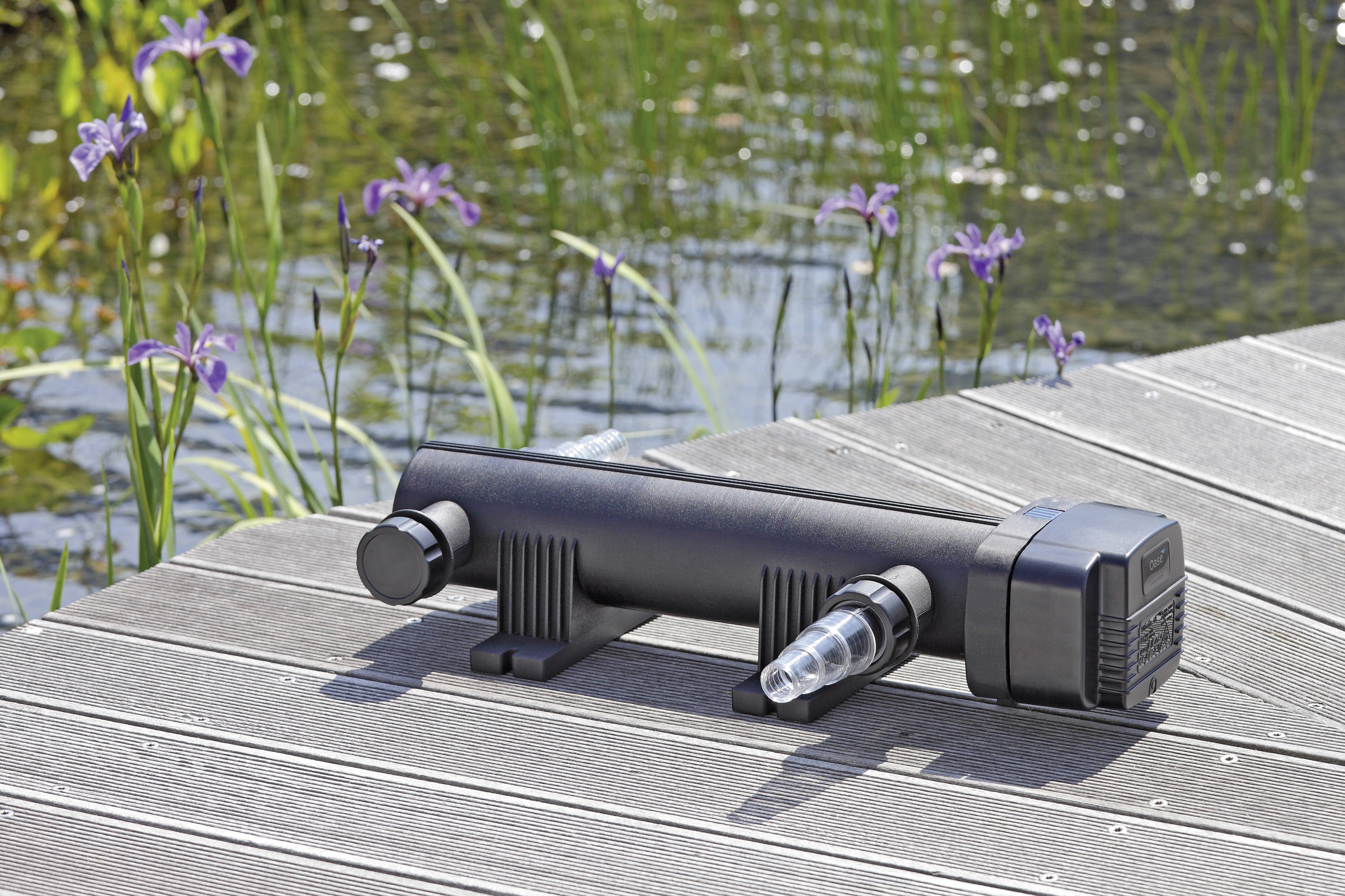 Schwarzes UV-Wasserfiltersystem auf einem Holzsteg an einem Teich mit lila Blumen und grünen Schilf im Hintergrund, bei Sonnenlicht.