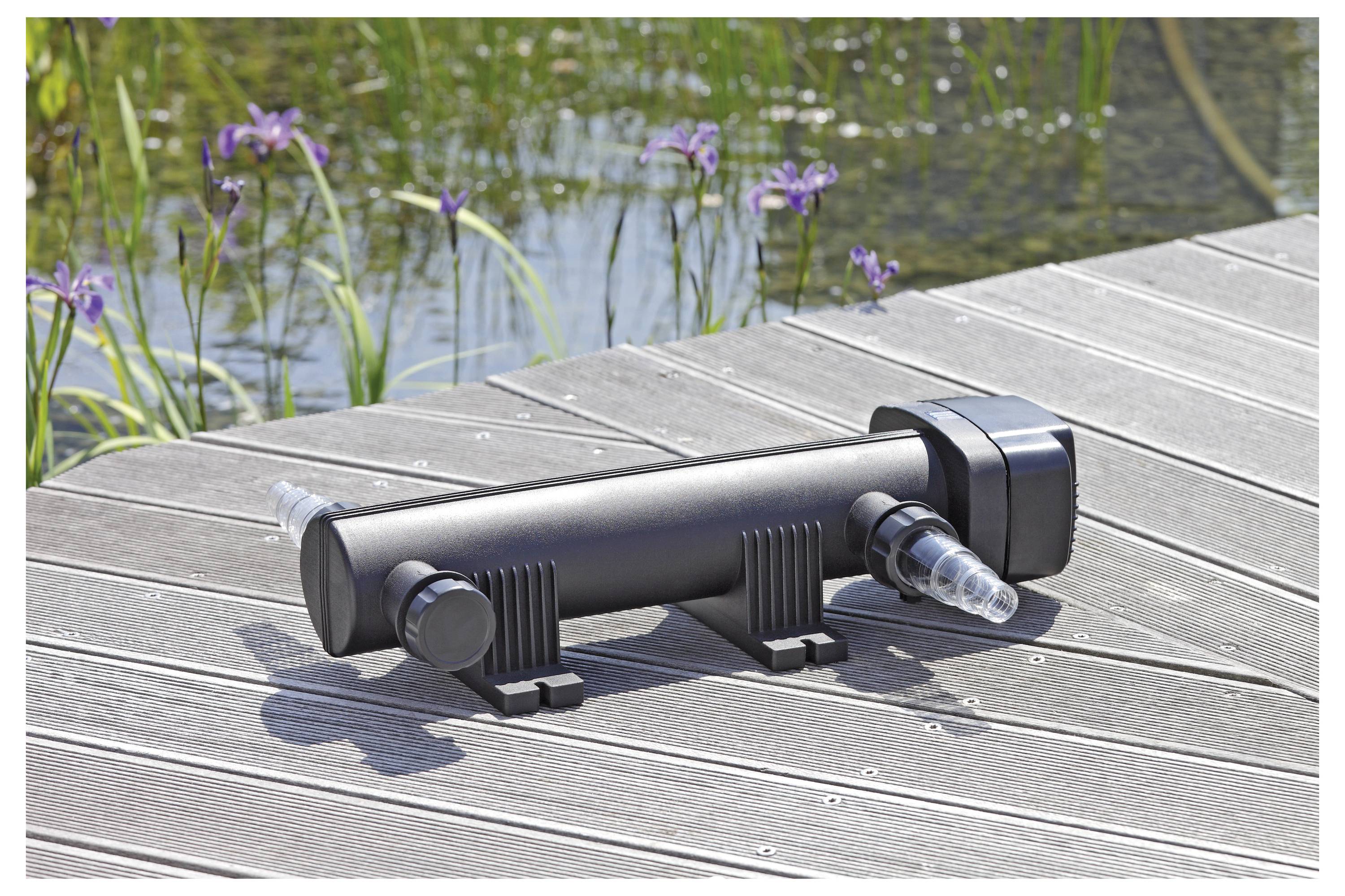 Ein schwarzer UV-Klärfilter, der auf einem Holzdeck neben einem Teich mit lila Blumen im Hintergrund platziert ist.