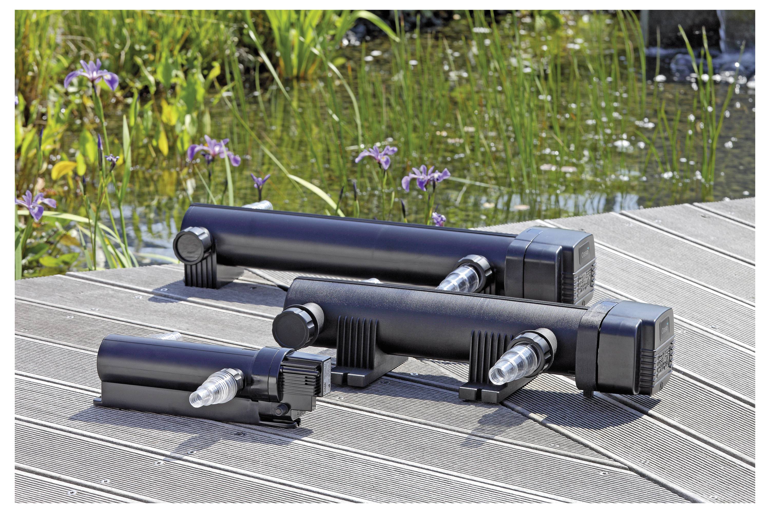 Drei schwarze UV-Wasserklärgeräte unterschiedlicher Größe sind auf einem Holzdeck neben einem Teich mit blühenden violetten Blumen platziert.