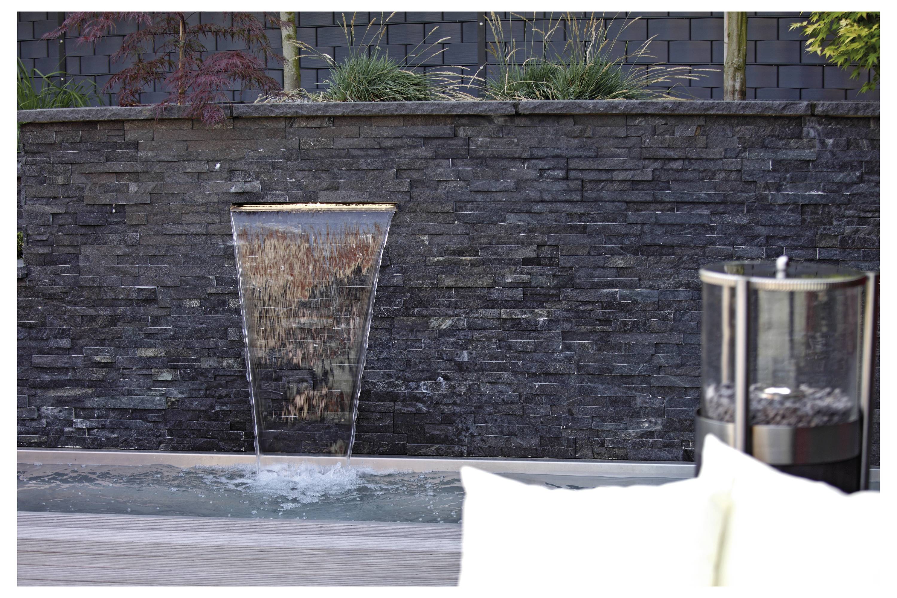 Ein dekorativer Wasserlauf an einer Steinmauer mit einem kleinen Wasserfall in einen schmalen Teich. Grüne Pflanzen befinden sich oberhalb der Mauer.