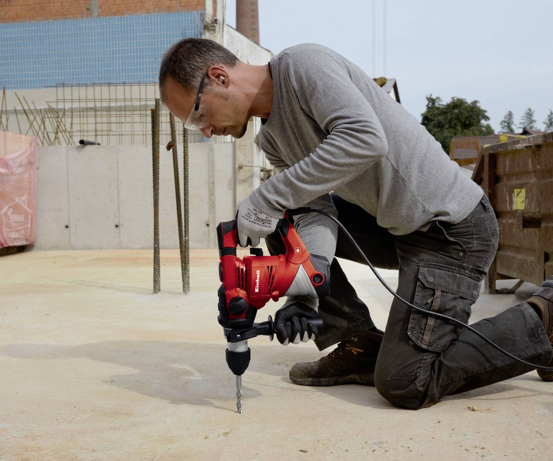 Arbeiter kniet auf einer Baustelle und benutzt eine rote Bohrmaschine auf Betonboden. Hintergrund zeigt Bauaktivitäten.