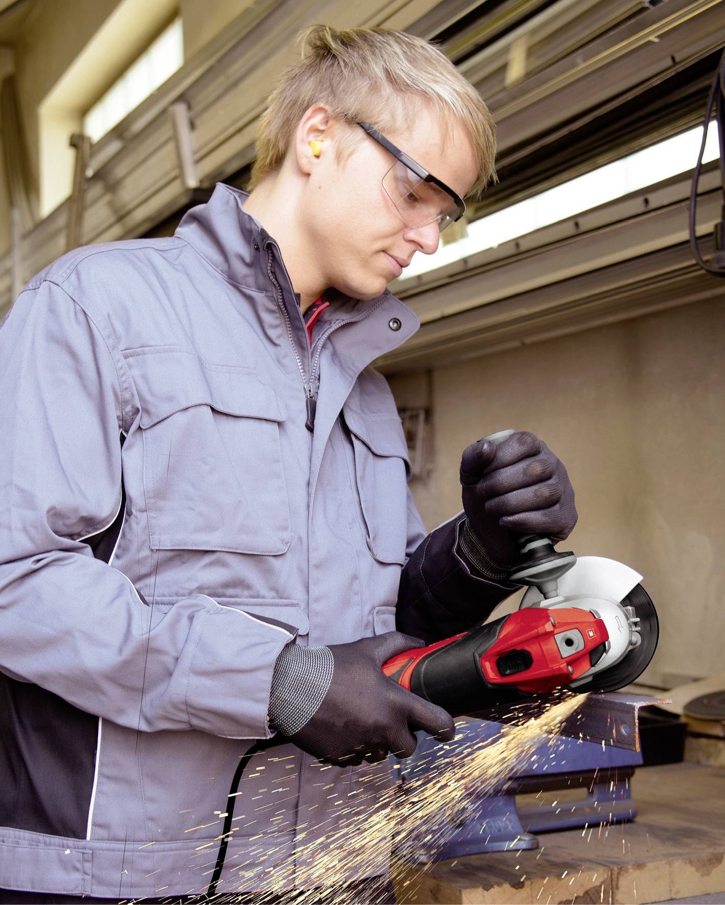 Ein Mann in Schutzbrille und Handschuhen schleift Metall mit einem Winkelschleifer in einer Werkstatt. Funken sprühen beim Schleifen.