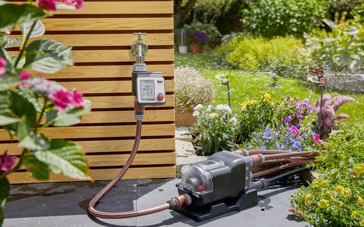Ein Gartenbewässerungssystem, das Pflanzen automatisch mit einem an einer Außenwand angeschlossenen Schlauch bewässert, umgeben von Blumen und Grünpflanzen.