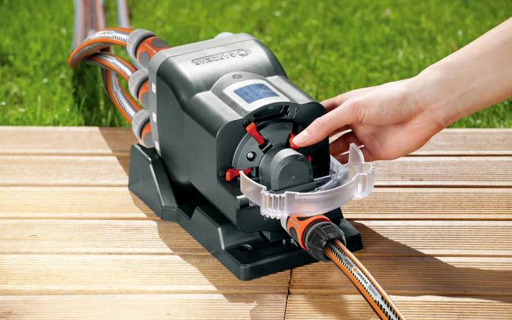 Eine Hand justiert einen Gartenschlauch-Timer auf einer Holzterrasse. Der Timer ist mit mehreren Schläuchen verbunden und zeigt die Wasserflusskontrolle in einer Gartenumgebung.