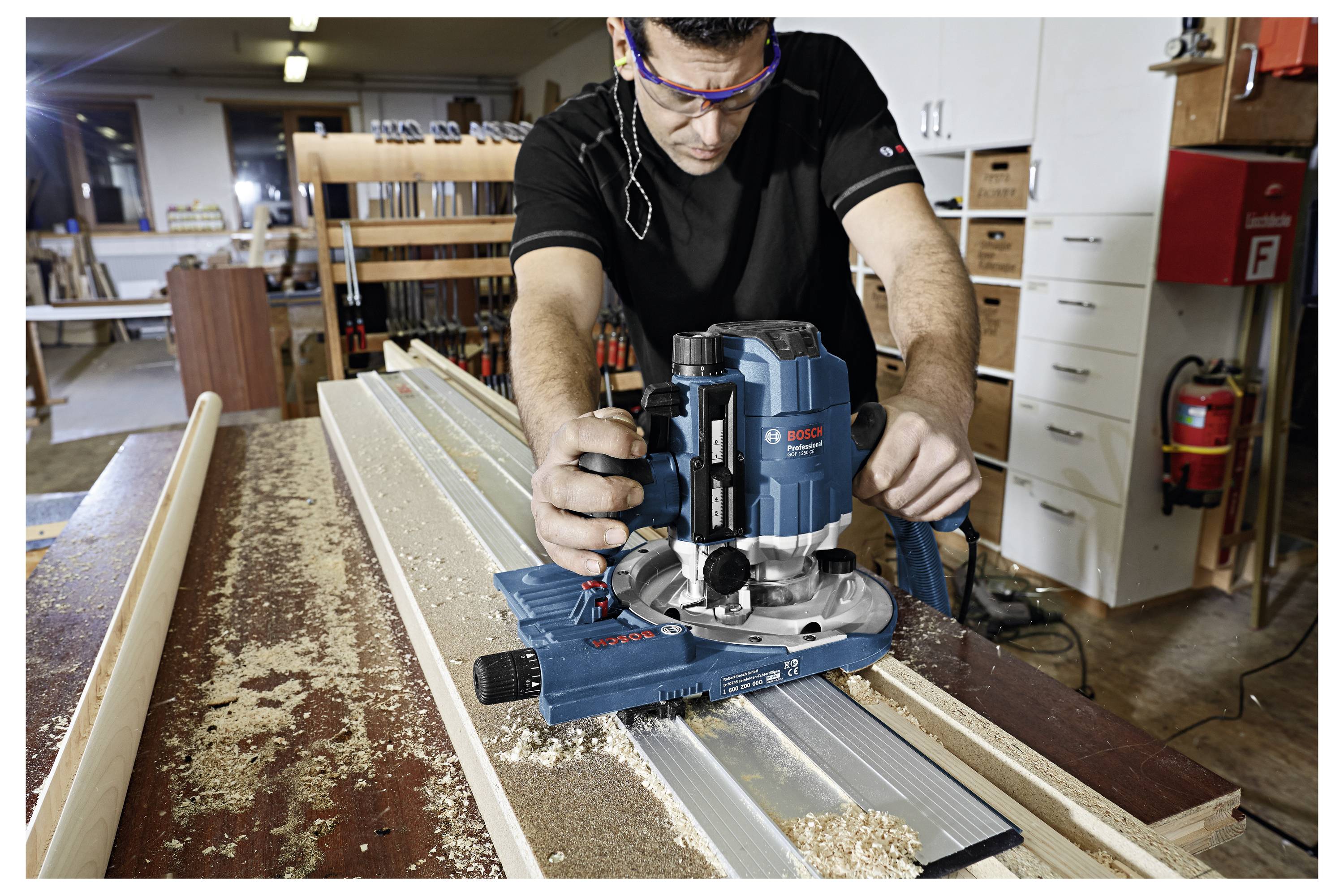 Handwerker nutzt Elektrowerkzeug zum Fräsen in Holzwerkstatt, um Holzleiste zu bearbeiten. Sägespäne auf Tisch, Regale im Hintergrund.