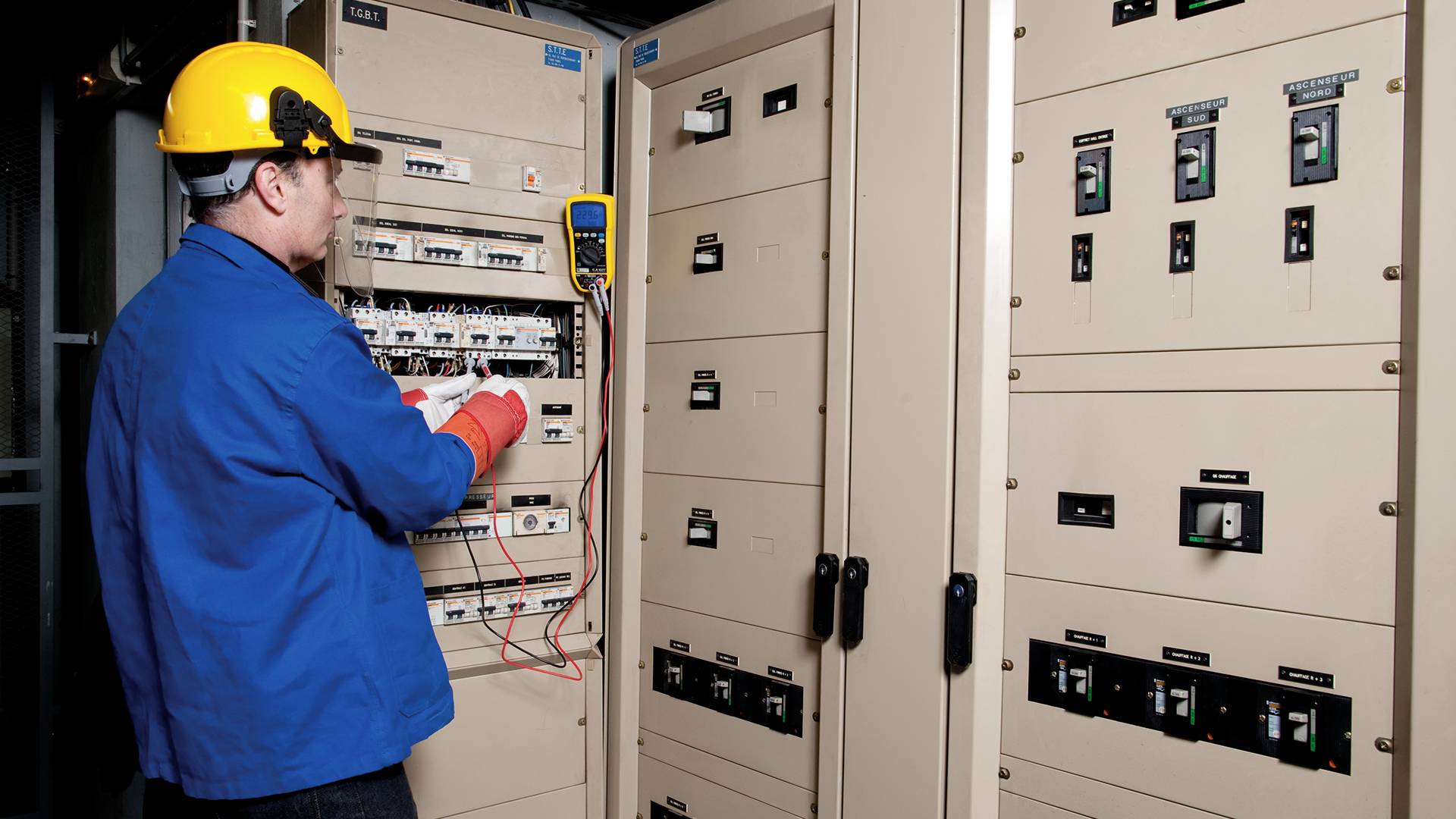 Ein Elektriker in blauer Arbeitskleidung und gelbem Helm prüft Schalttafeln mit einem Multimeter in einem Kontrollraum.