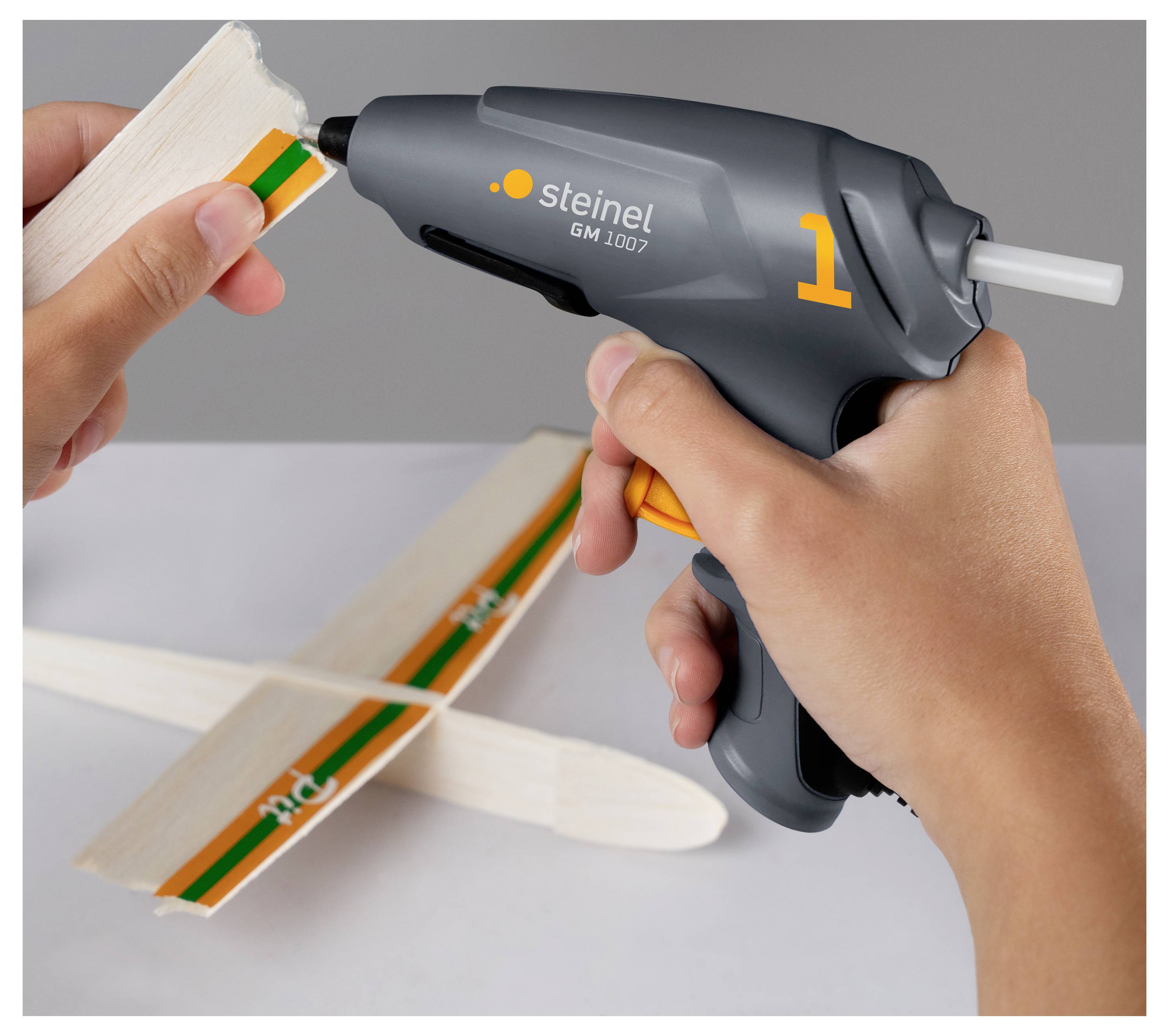 Hände, die eine Heißklebepistole verwenden, um Kleber auf das Ende eines Holzbastelstabs an einem kleinen Modellflugzeug aufzutragen.