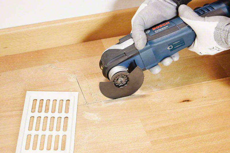 Eine Person schneidet mit einem oszillierenden Werkzeug ein rechteckiges Stück aus einem Holzboden neben einem weißen Lüftungsgitter.