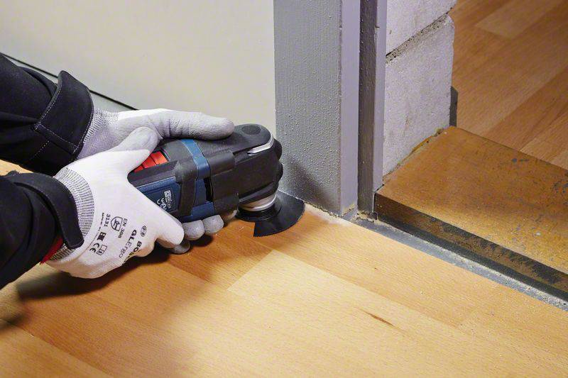Eine Person schneidet mit einem Multifunktionswerkzeug einen Holzboden an der Kante zur Türschwelle. Sie trägt Arbeitshandschuhe.