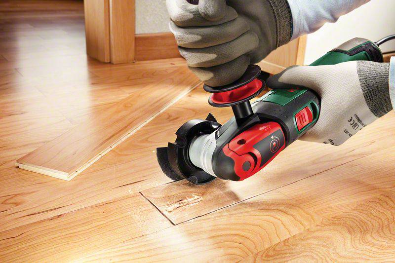 Eine Person sägt ein rechteckiges Stück aus einem Holzfußboden mit einem handgehaltenen Elektrowerkzeug. Holzspäne sind sichtbar.