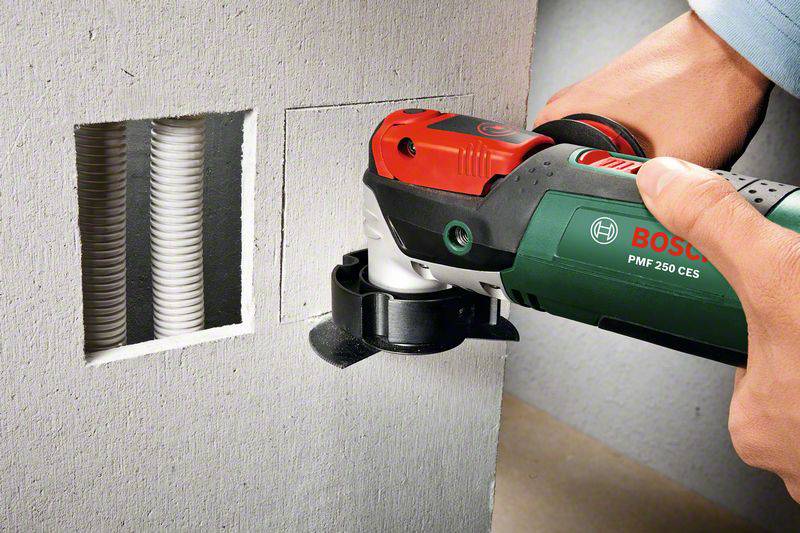 Eine Person verwendet ein grünes Elektrowerkzeug, um eine Aussparung in einer Wand zu schneiden. Rechts ist ein Kabelschacht sichtbar.