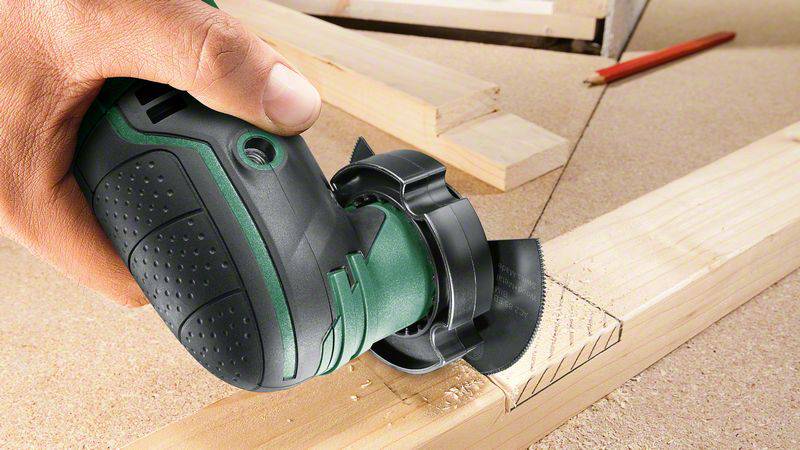 Eine Person sägt Holz mit einem grünen Elektrowerkzeug. Ein Bleistift liegt im Hintergrund.