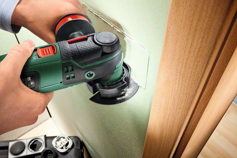 Eine Person schneidet mit einem elektrischen Multifunktionswerkzeug ein rechteckiges Loch in eine grüne Wand neben einem Holztürrahmen.