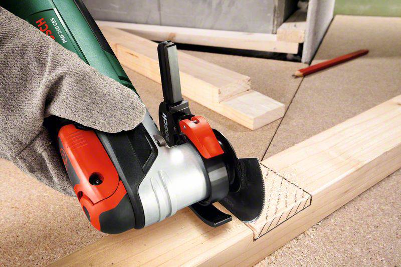 Eine Hand mit Arbeitshandschuh hält einen elektrischen Winkelschleifer, der auf ein Stück Holz mit markierten Schnittlinien gerichtet ist.