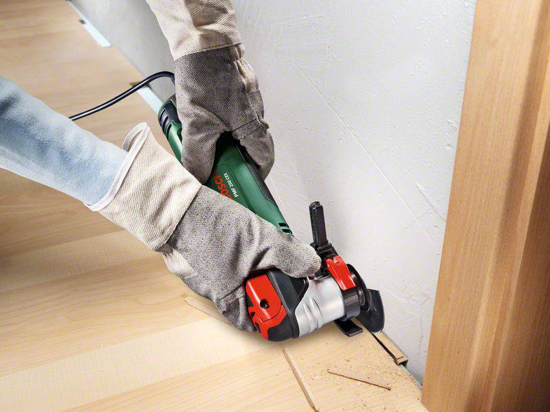 Eine Person trägt Handschuhe und verwendet ein oszillierendes Multifunktionswerkzeug, um Holz entlang einer Wand präzise zu schneiden.