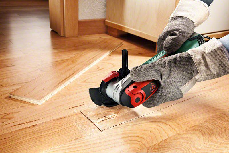 Person schneidet Holzboden mit einer elektrischen Säge in einem Wohnraum. Schutzhandschuhe werden getragen.
