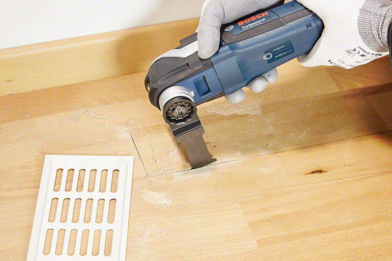 Eine Person verwendet ein Elektrowerkzeug, um ein rechteckiges Loch in eine Holzplatte zu schneiden. Ein Lüftungsgitter liegt daneben.