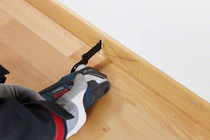Eine Person nutzt ein Elektrowerkzeug, um eine Holzleiste am Boden zu bearbeiten. Die Leiste hat Bleistiftmarkierungen als Schnittlinien.