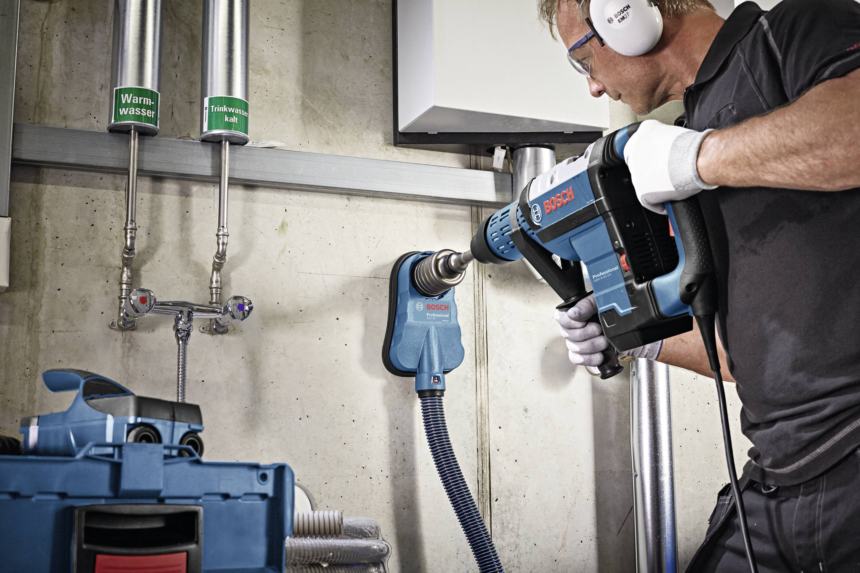 Ein Handwerker arbeitet mit einem Bohrhammer an einer Betonwand. Neben ihm sind Warmwasser- und Kaltwasserleitungen sichtbar.