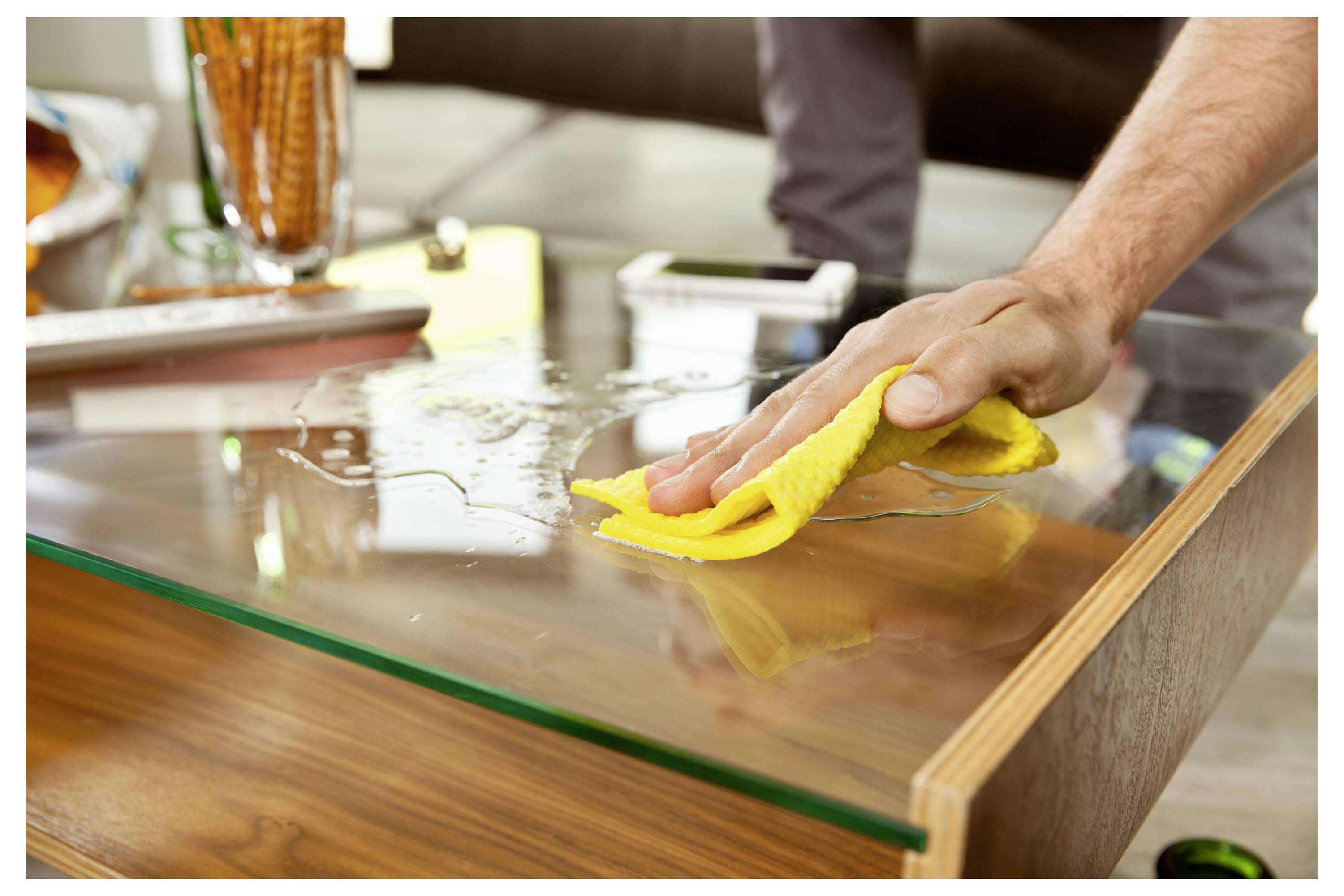 Person, die einen Glaskaffeetisch mit einem gelben Tuch abwischt, reinigt eine verschüttete Flüssigkeit auf einem Holztisch