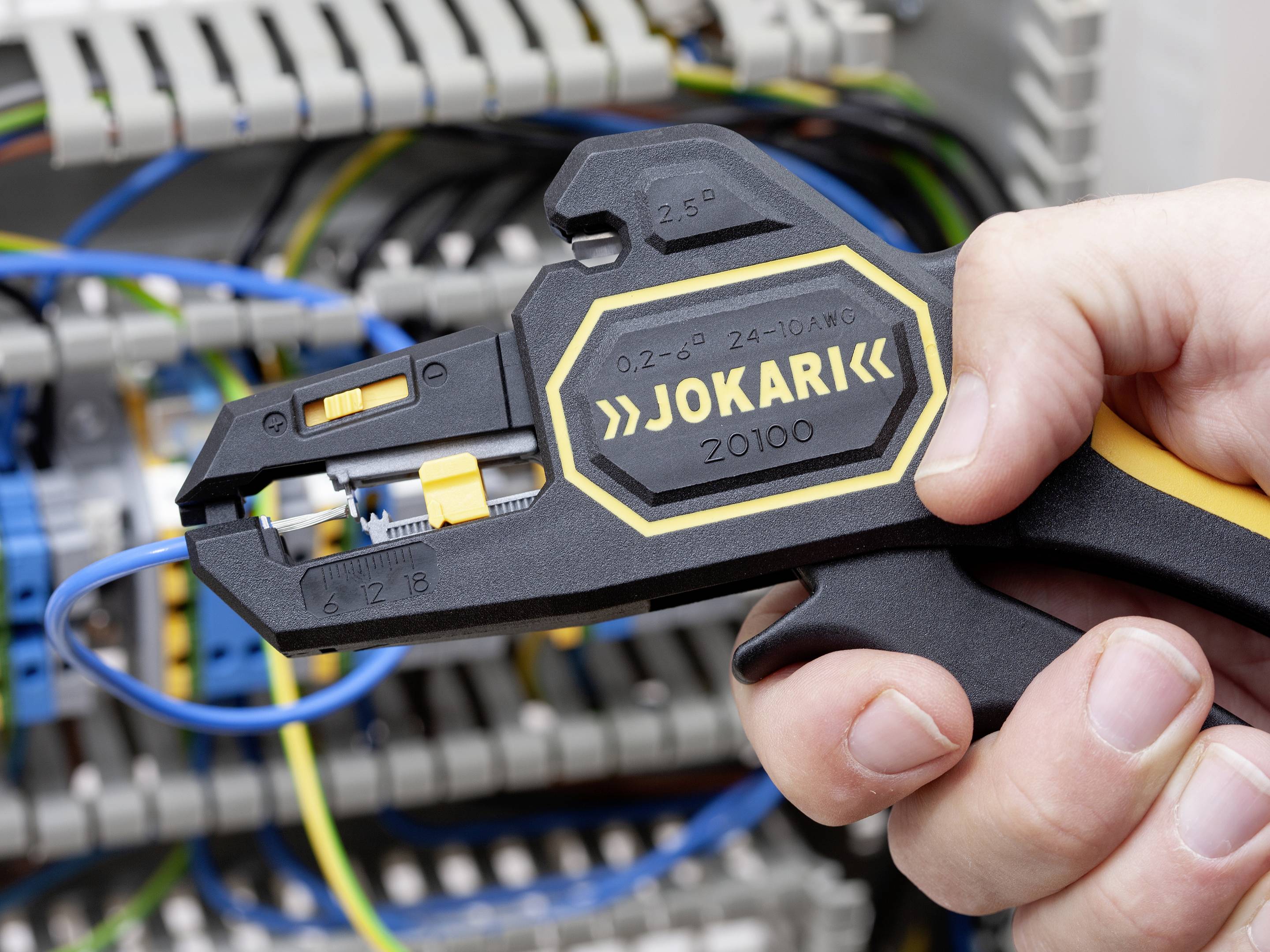 Eine Person benutzt einen Abisolierer, um ein Kabel in einer Schalttafel zu bearbeiten. Der Abisolierer ist der Marke 'Jokari'.
