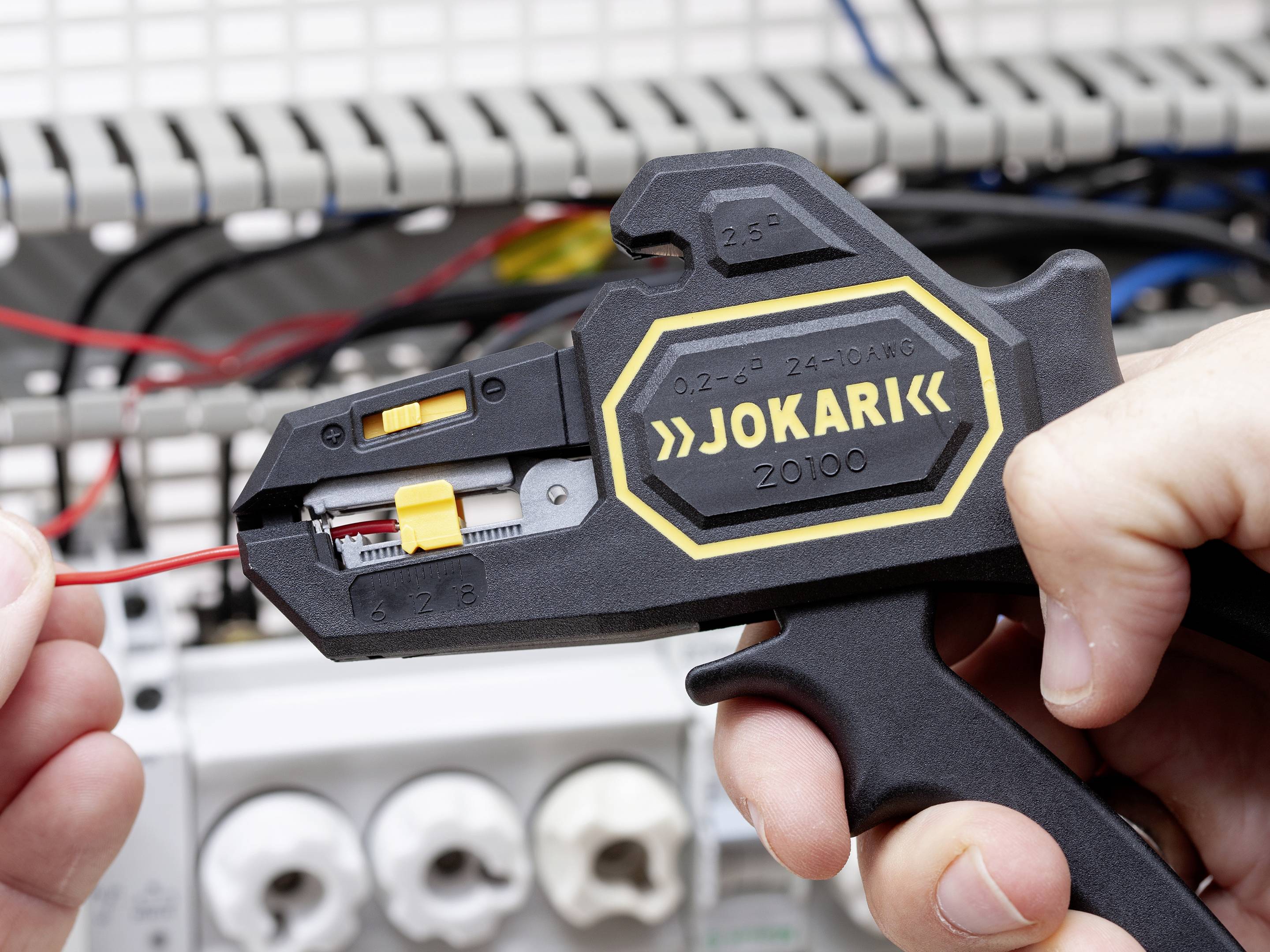 Eine Person benutzt einen Jokari-Drahtschneider, um einen roten Draht in einem Schaltschrank abisolieren.