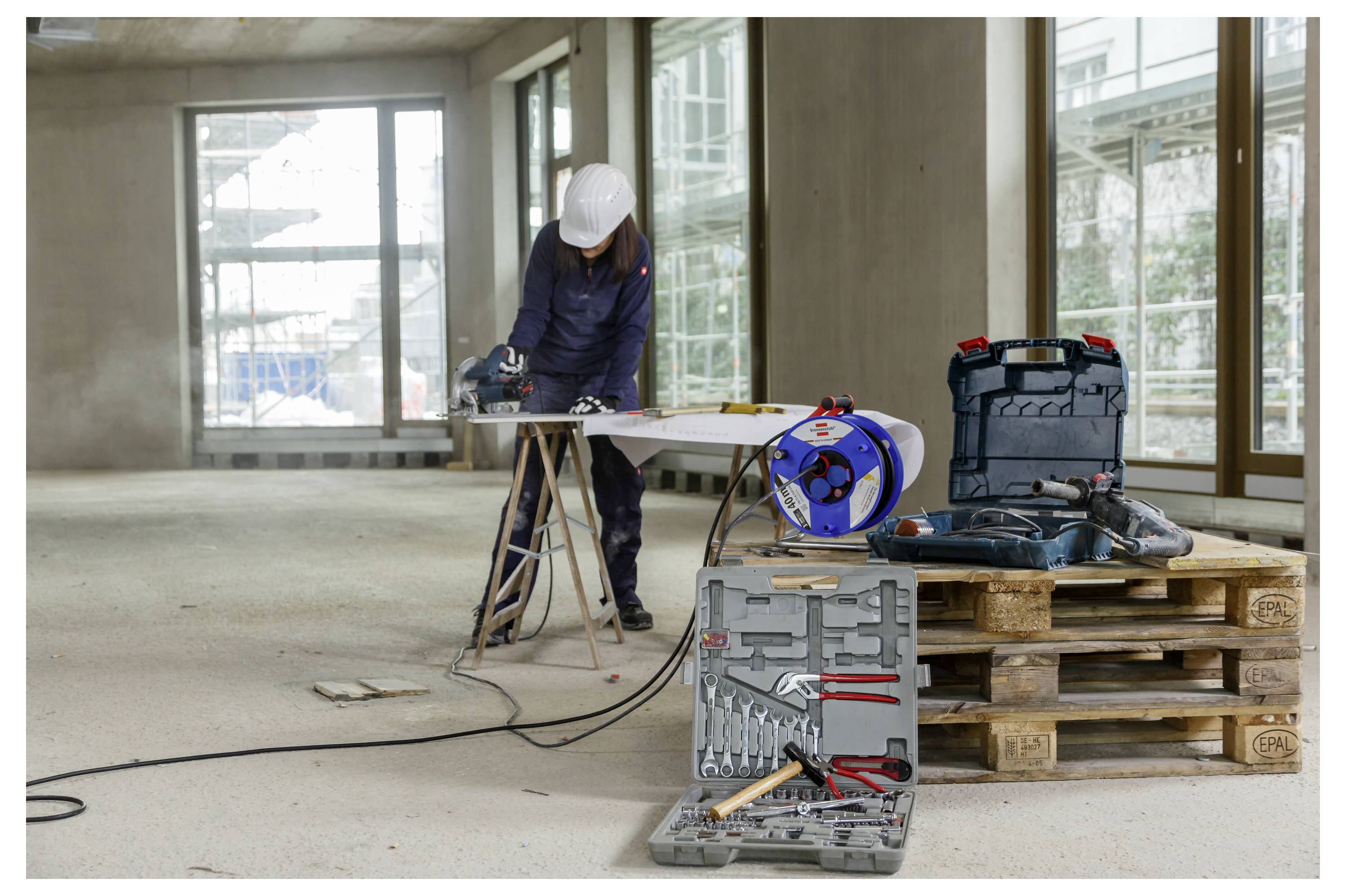 A construction worker uses a saw on a wooden board inside an unfinished building, surrounded by tools and equipment on pallets.