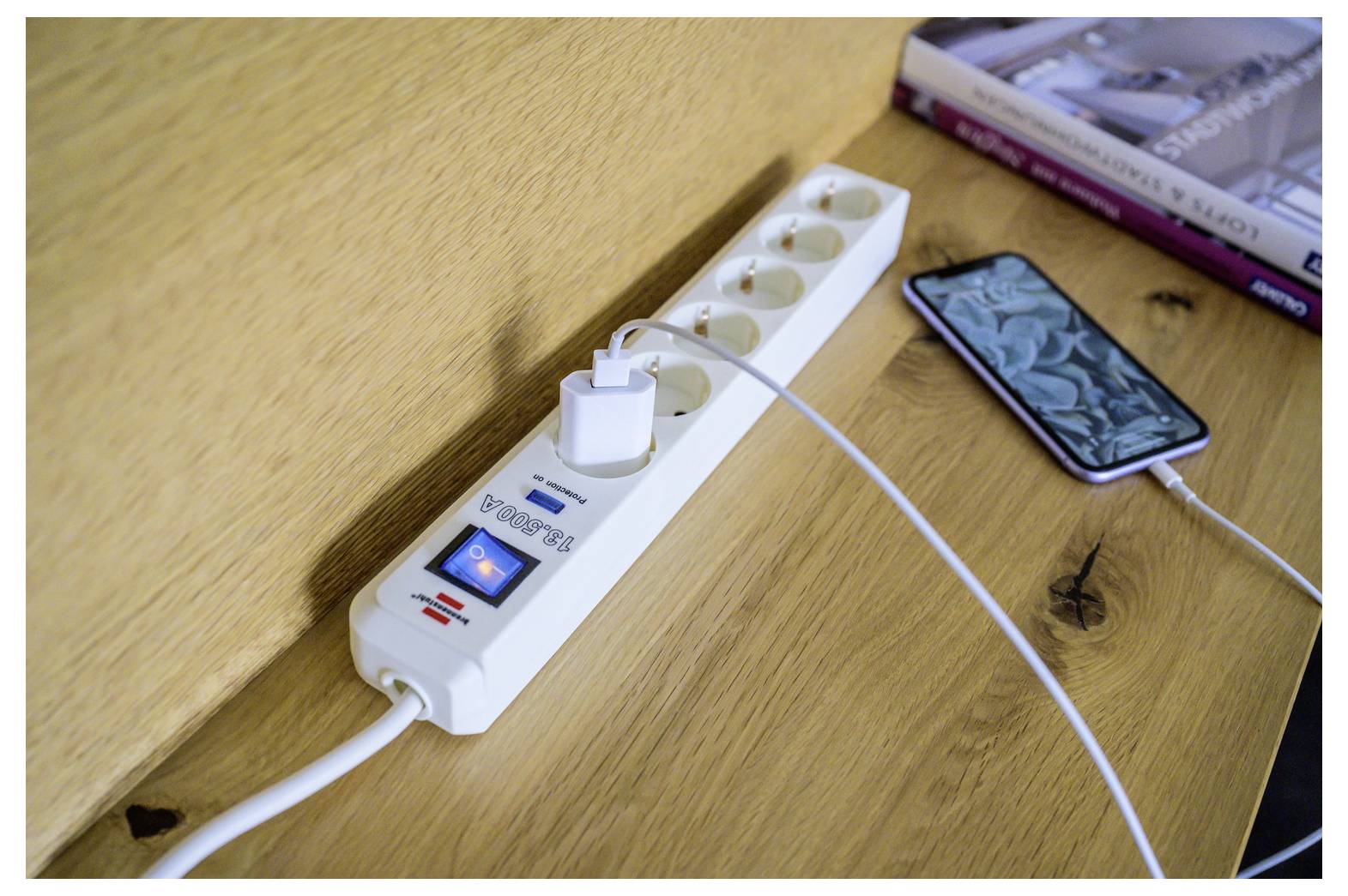 Stromleiste mit eingecktem Telefonladegerät auf einem Holzschreibtisch, neben einem Smartphone und Büchern, die ein Geräteladeset zeigen.