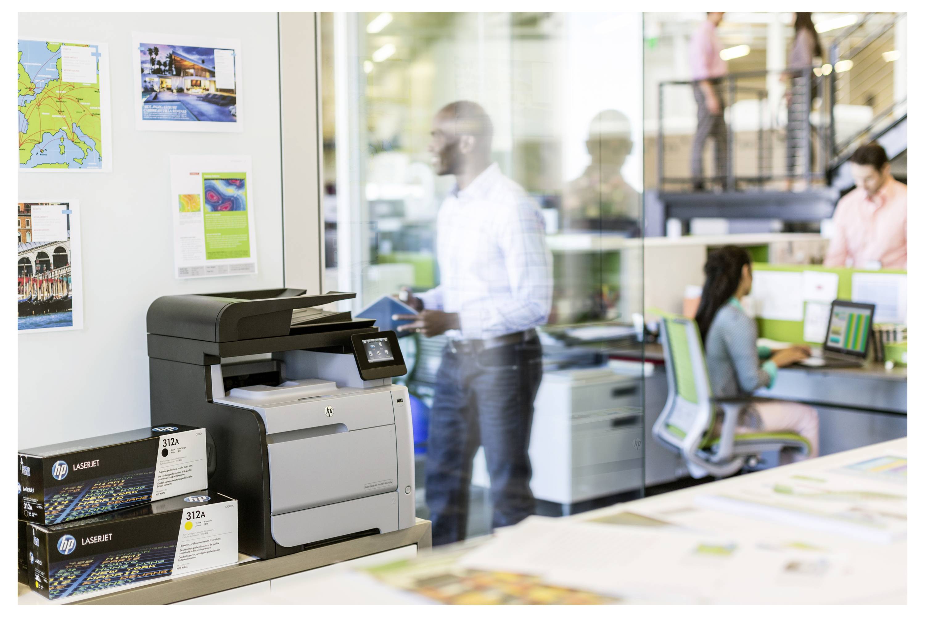 Büro mit einem großen LaserJet-Drucker im Vordergrund, während Kollegen in einem Arbeitsbereich mit Glaswänden dahinter sitzen und stehen.